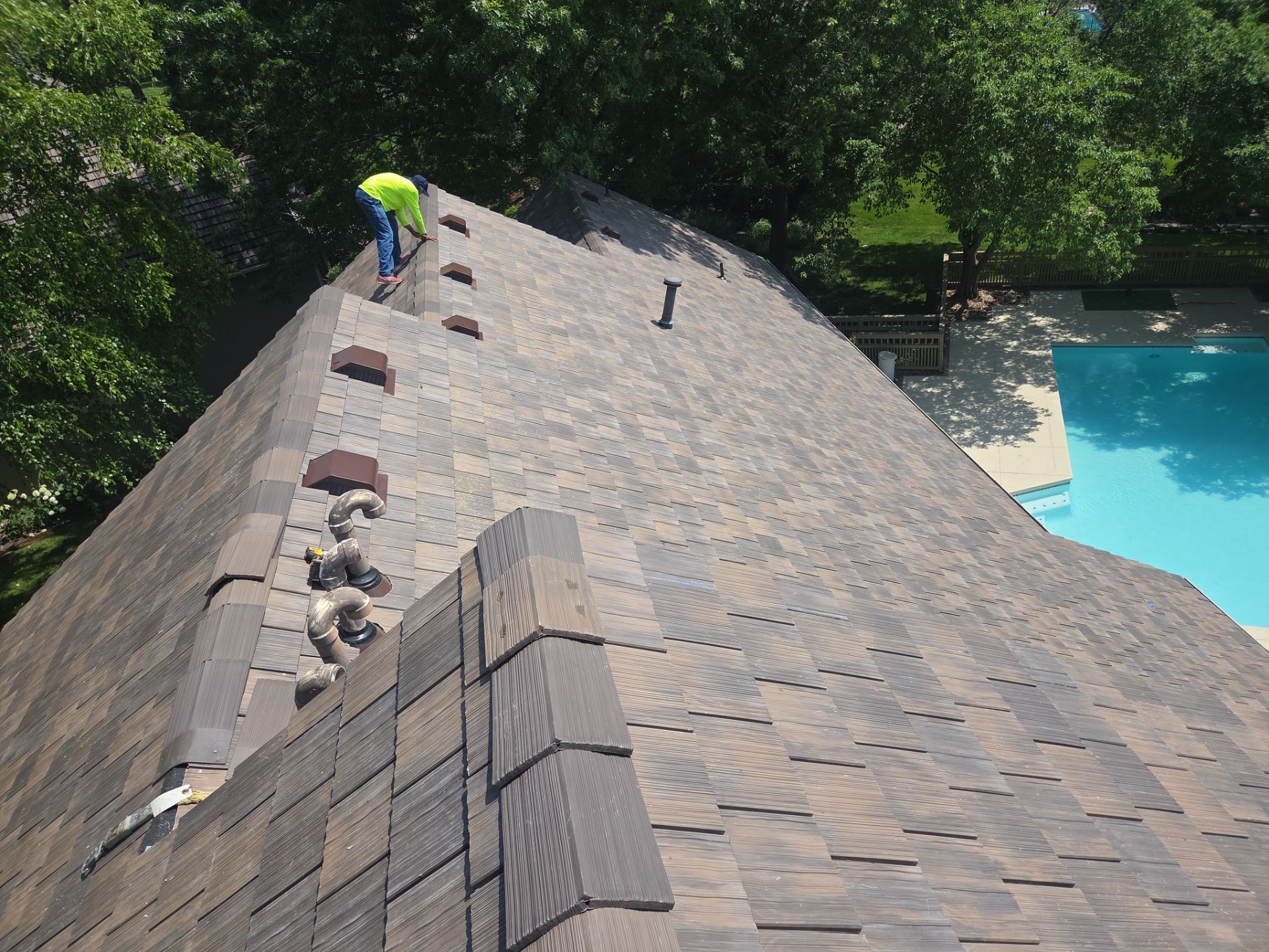Roofer repairing damaged asphalt shingles on a house roof near a pool.