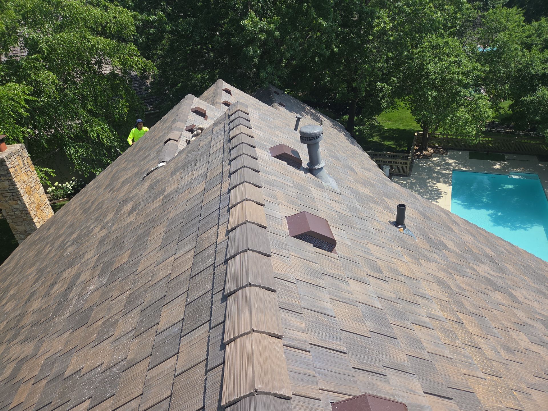 View of a brown shingle roof with a chimney and vents, with a pool visible in the background.