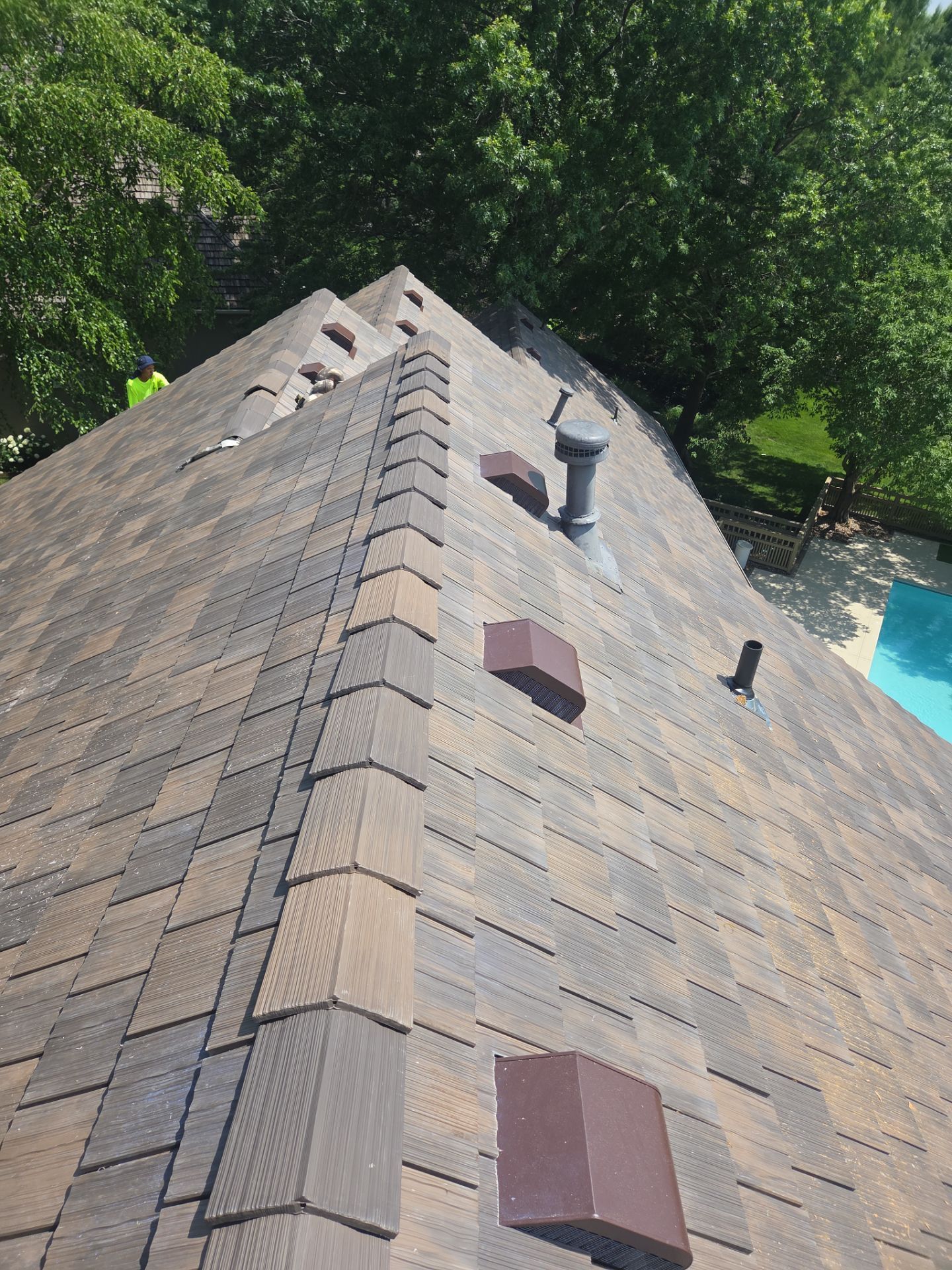 A roof covered in brown shingles, viewed from above. A chimney and vents are visible; trees and a pool are in the background.