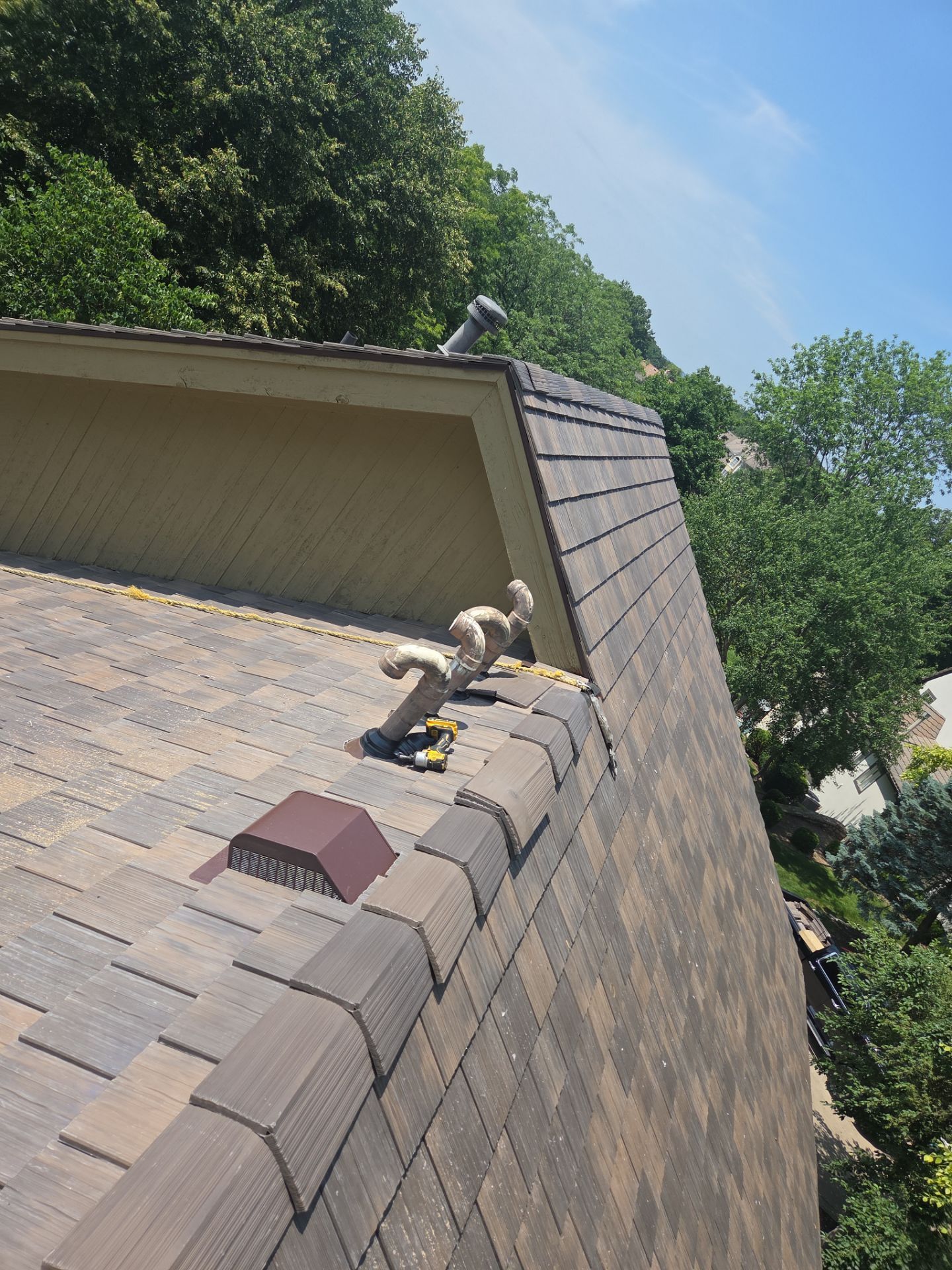 Rooftop with shingles, tools, and vents against a backdrop of green trees and a partly cloudy sky.