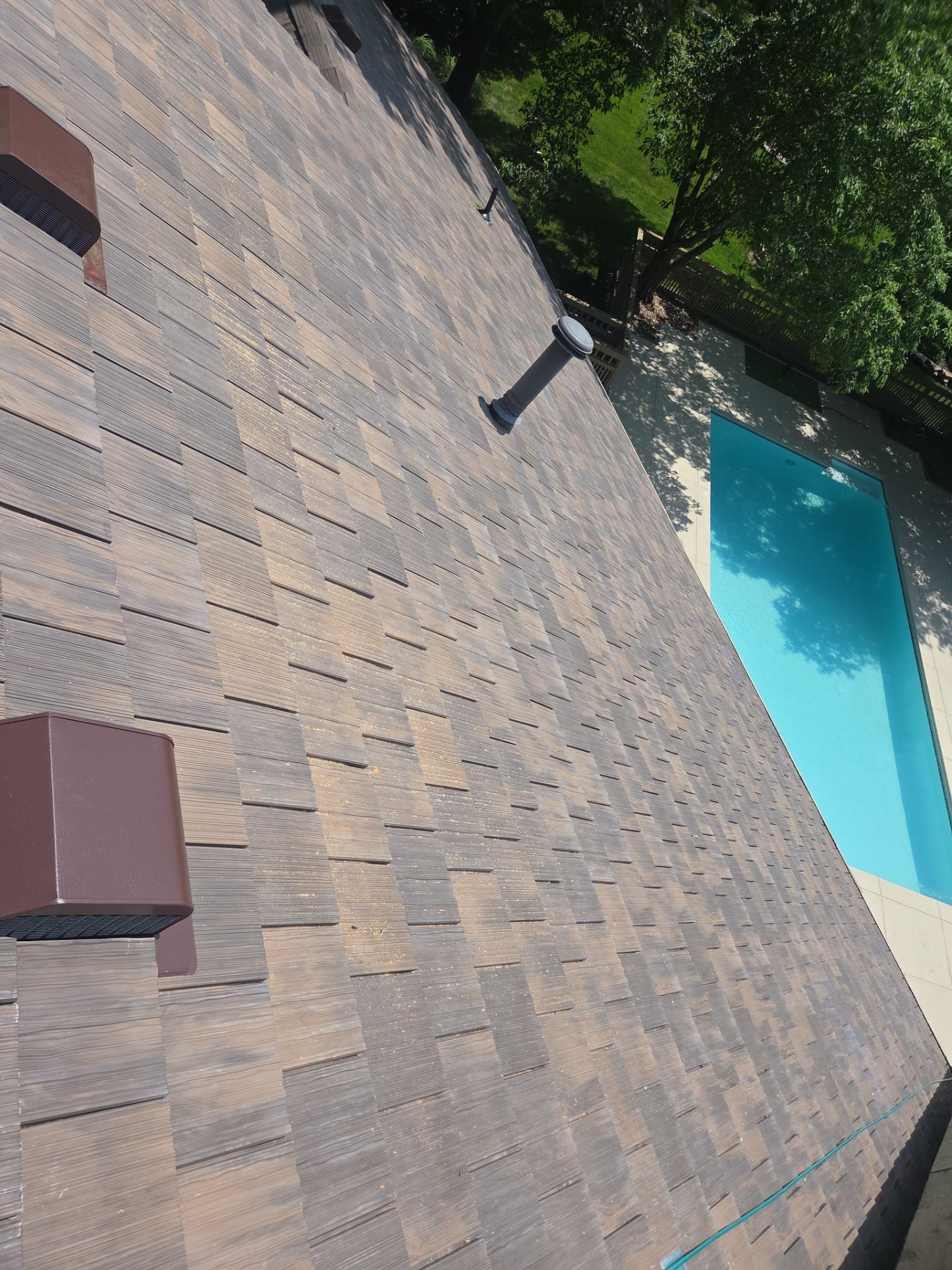 Brown shingled roof with two dark brown vents, a pool on the right, and some trees.