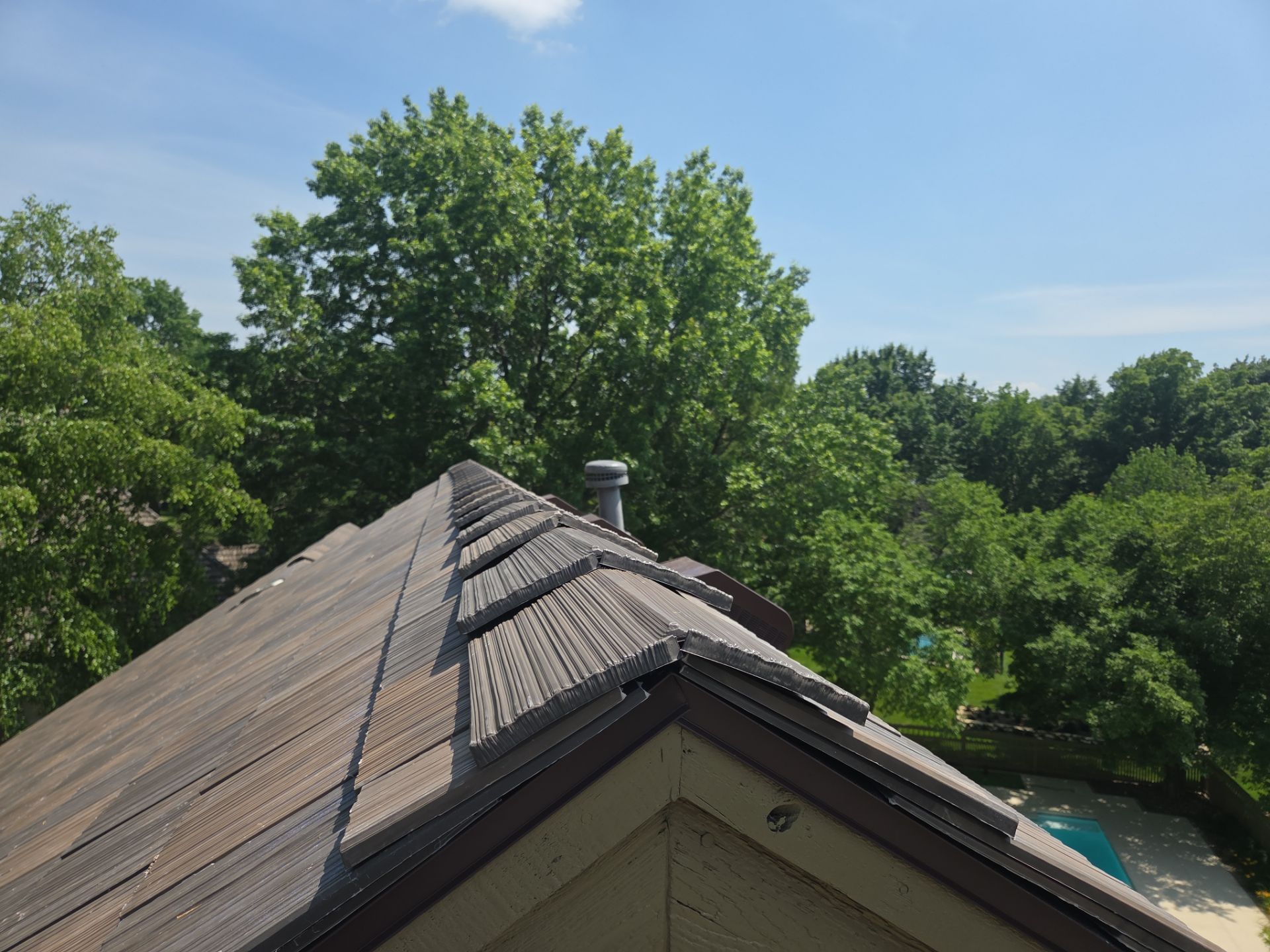 View from a roof: weathered shingles, chimney, lush green trees, and a pool under a blue sky.