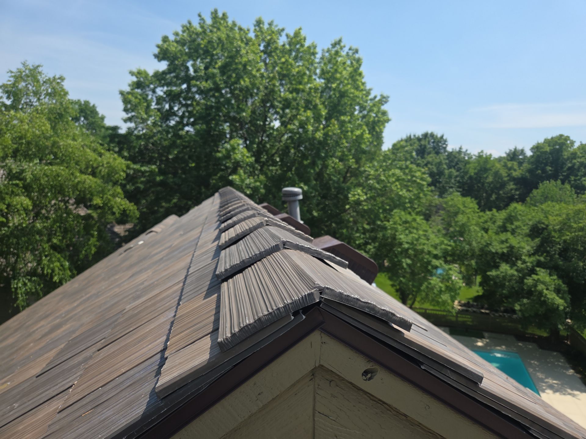 Damaged roof shingles on a residential home, with a background of green trees and a pool.