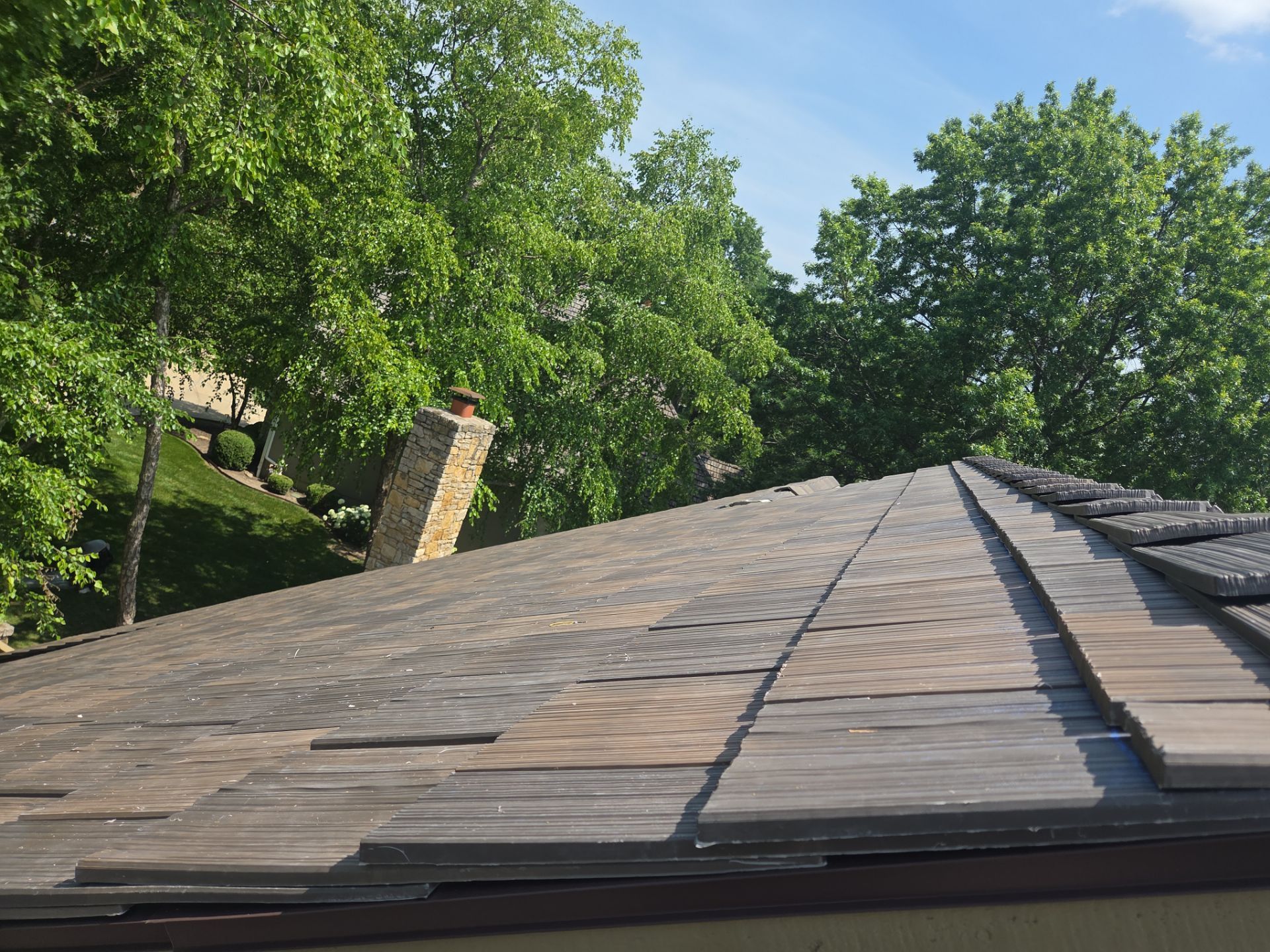 A weathered roof with missing shingles; trees and chimney in the background on a sunny day.