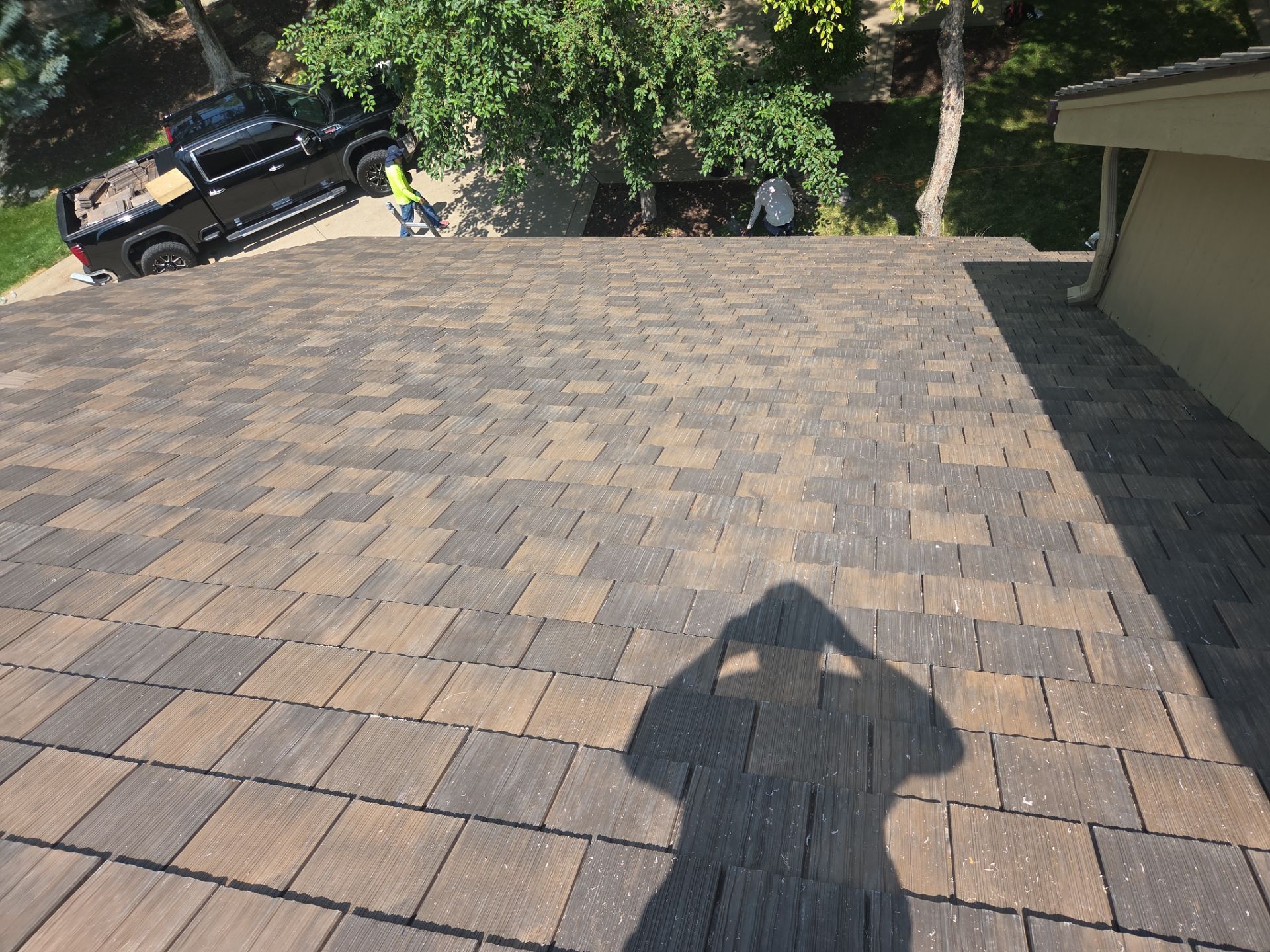 A person's shadow on a brown and tan shingle roof. A black truck is parked nearby.