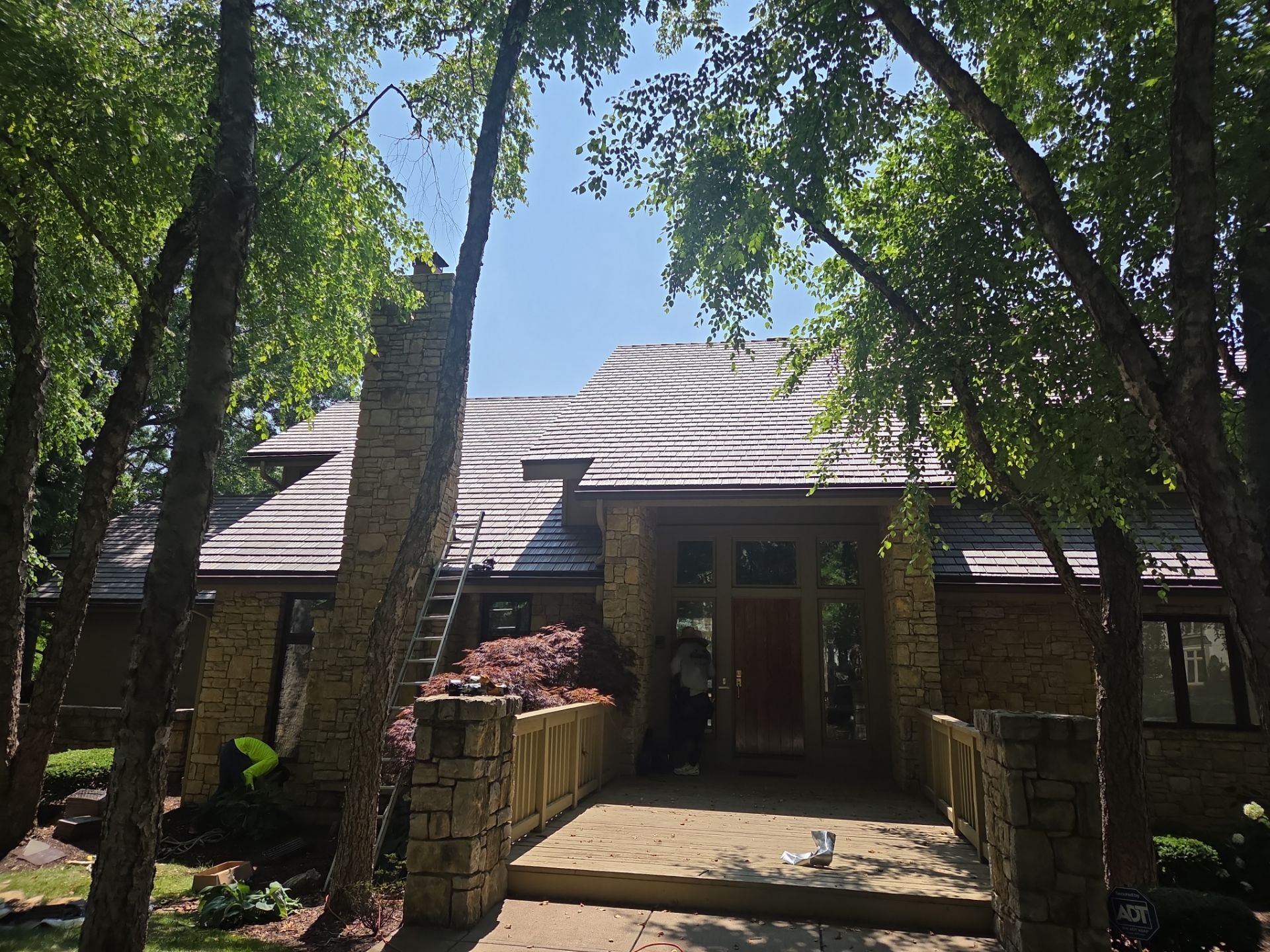 House with stone facade, wooden roof, and walkway; surrounded by trees under a blue sky.