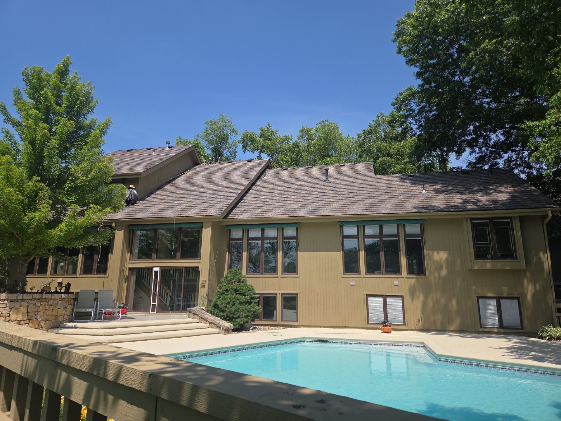 Tan house with pool and brown roof surrounded by trees under a blue sky.
