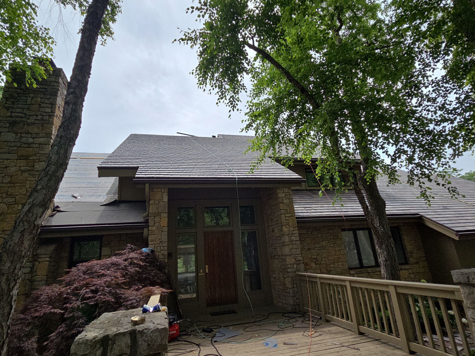 House with stone facade and damaged roof under overcast sky. Trees surround the entrance.
