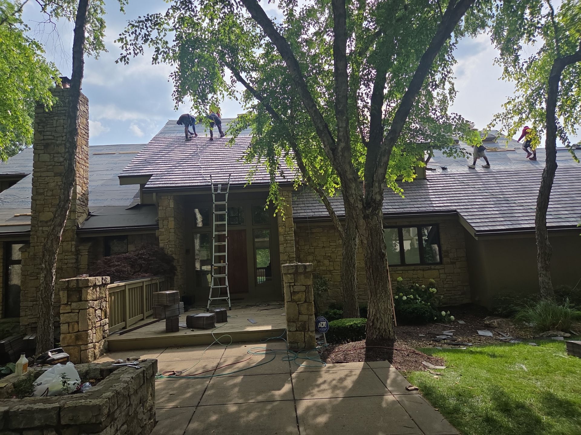 Roofers replace a roof on a house, with trees in the foreground.