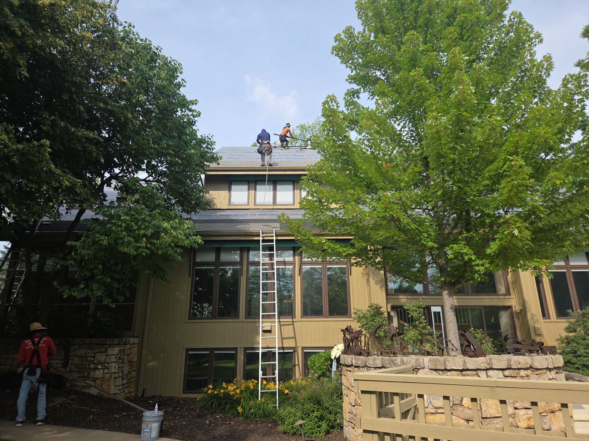 Roofers working on a two-story house roof, trees on either side, sunny day.