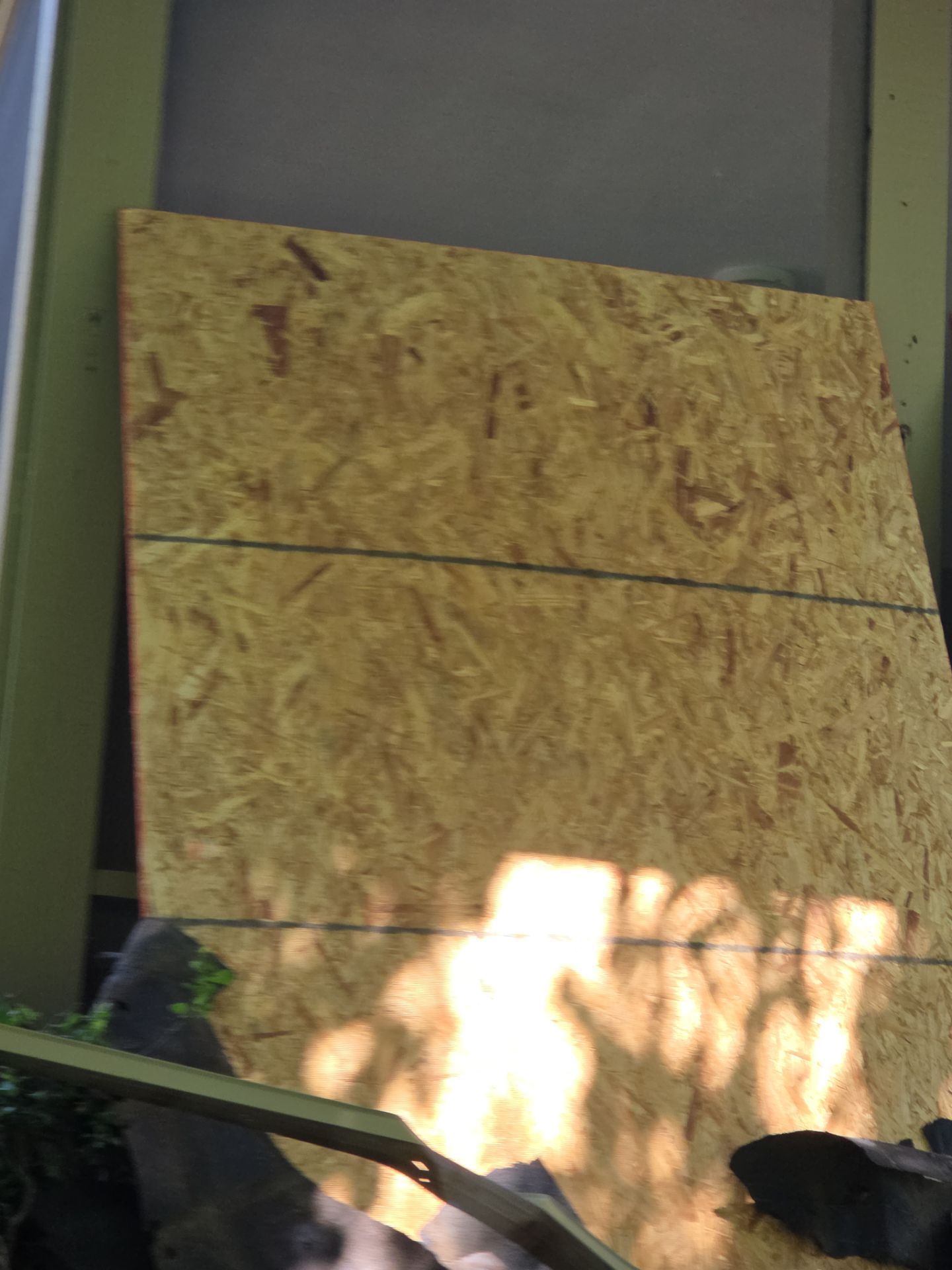 A piece of plywood leaning against a light green structure; sunlight casts shadows.
