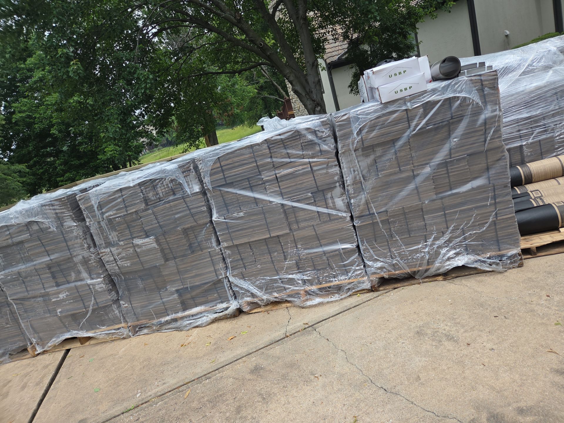 Pallets of gray pavers wrapped in plastic, outdoors near a tree and building.