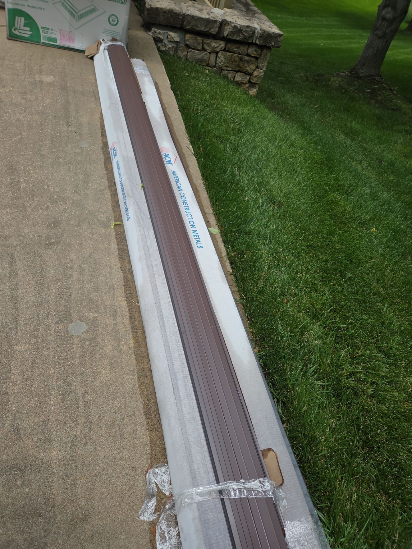 Long, brown gutter guard lying on a concrete surface, next to green grass.