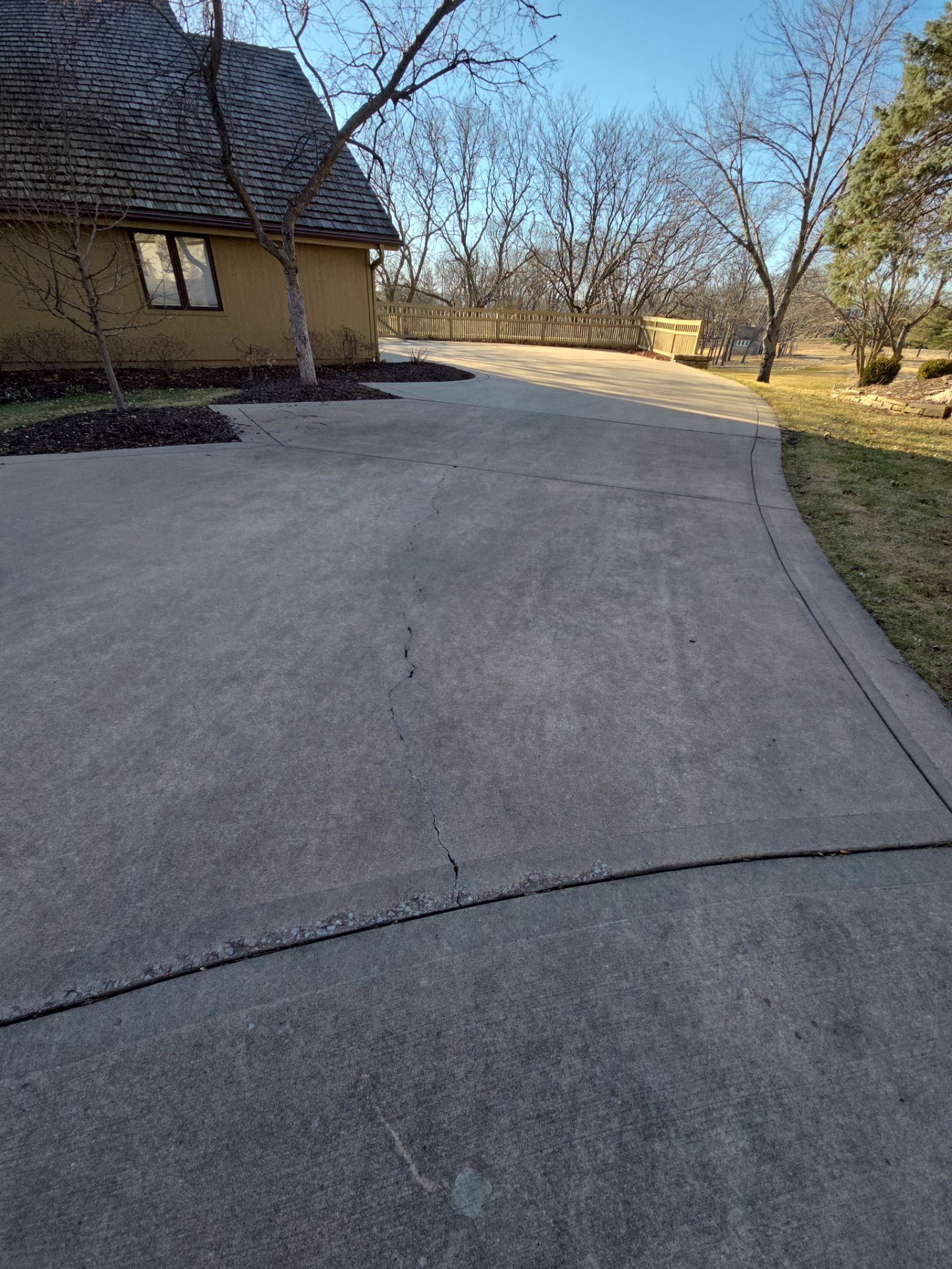 Driveway curves toward a tan house with a brown shingle roof. Trees and grass line the side.