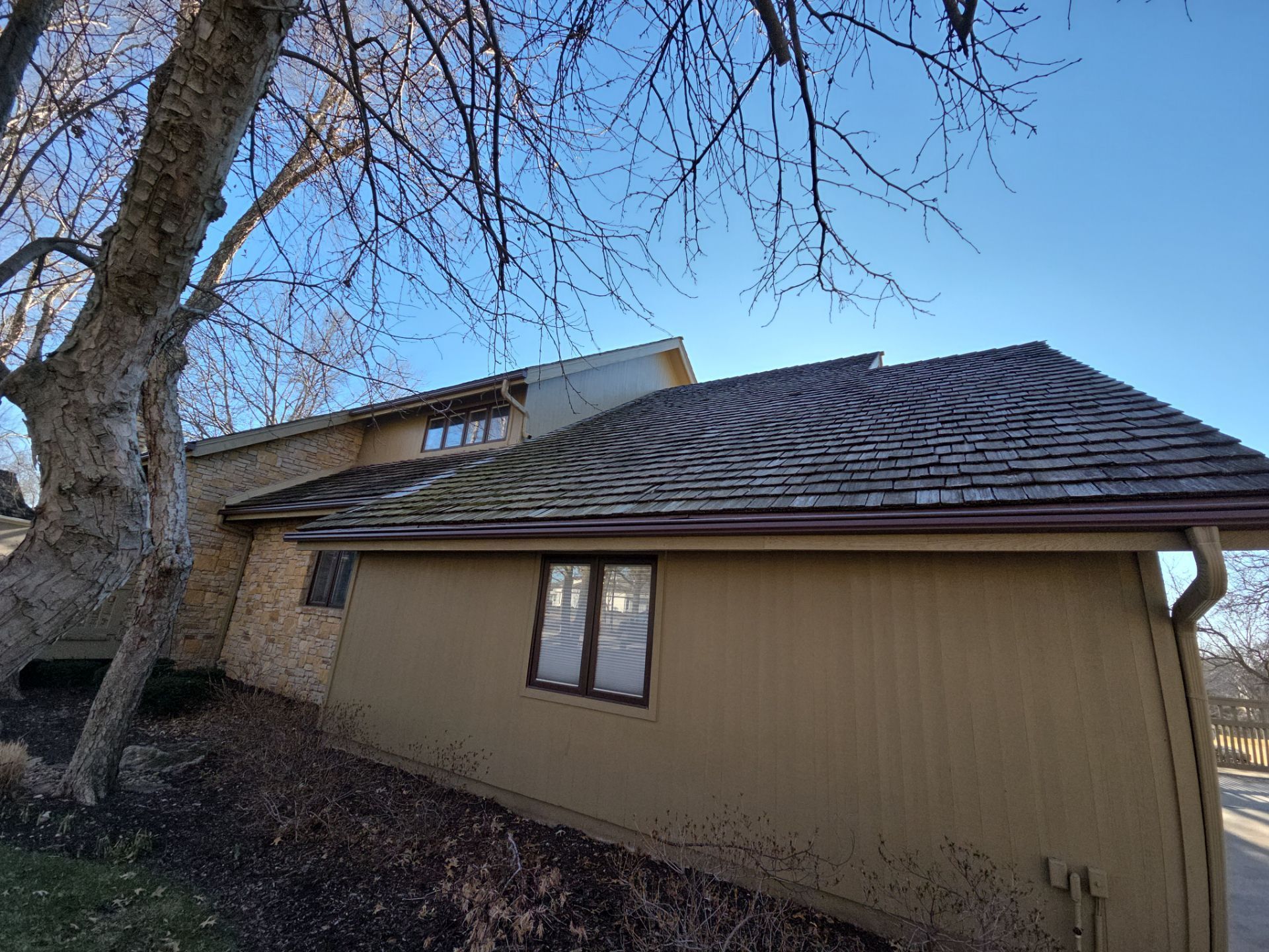 Beige house with brown roof and bare tree branches against a clear blue sky.