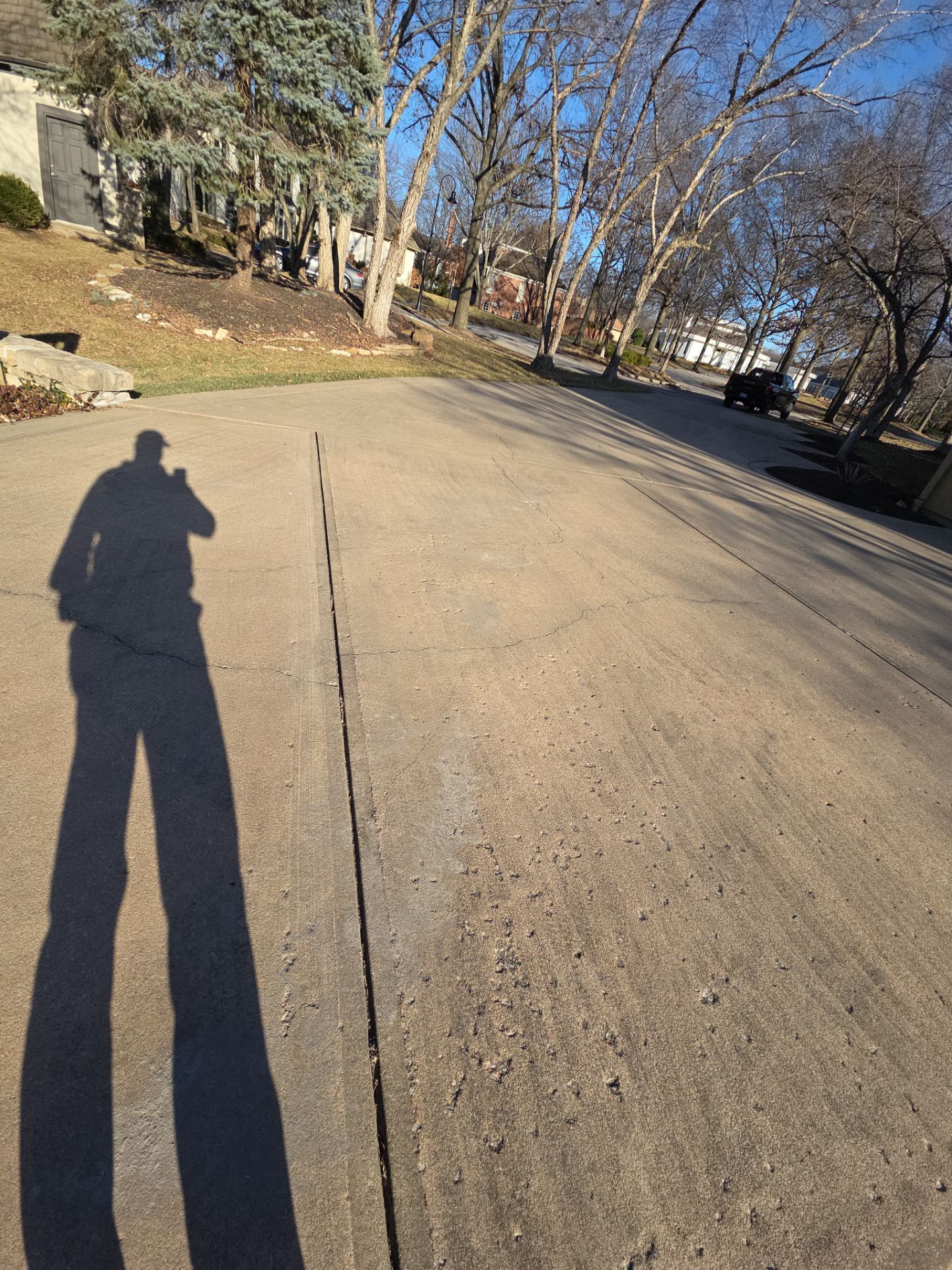 Person's shadow on a driveway; sunny day. Trees and homes visible in the background.