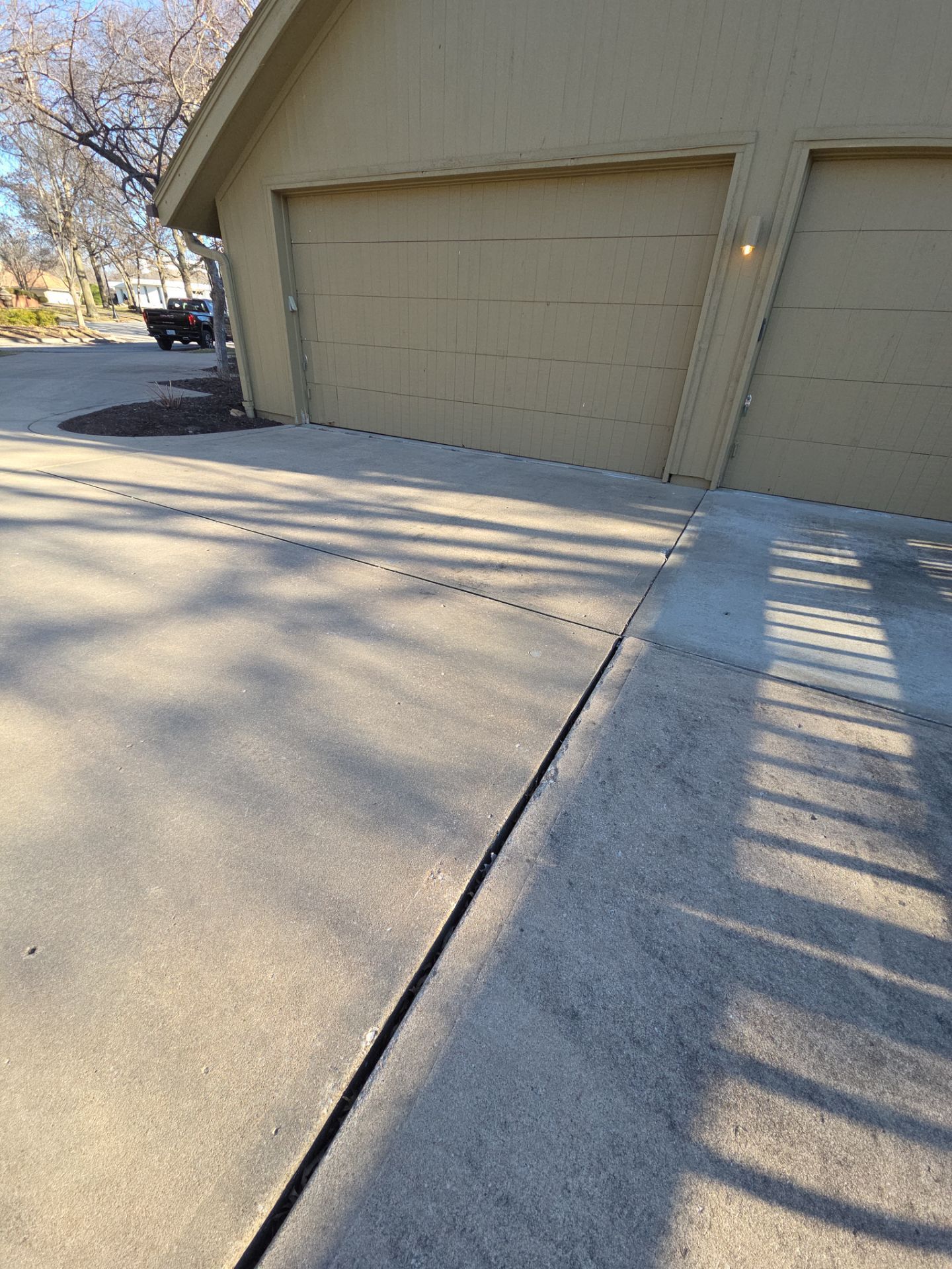 Concrete driveway leading up to a beige garage with two doors; long shadows from a tree.