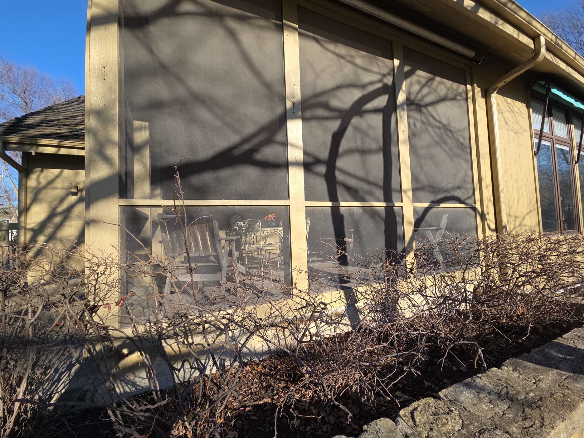 Screened-in porch with bare bushes in foreground; sunny day.