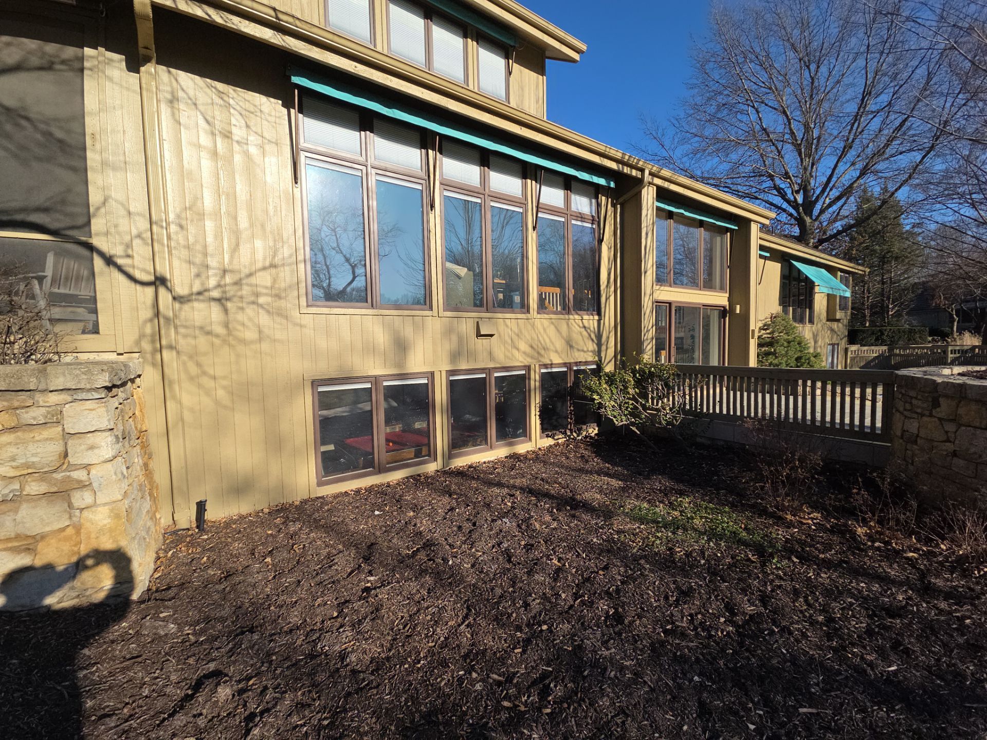 Tan house with large windows, brown mulch, and a wooden railing on a sunny day.