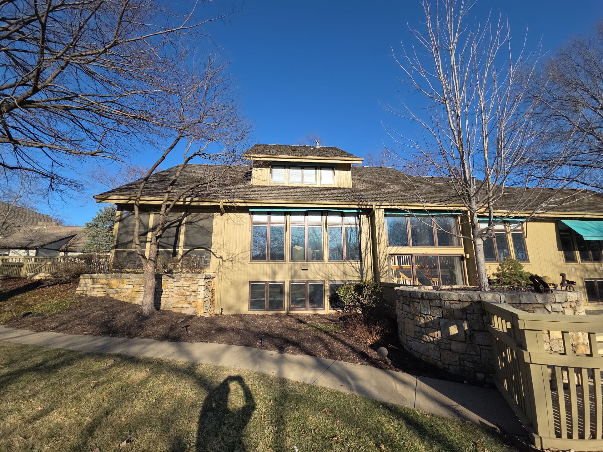 Tan house with a weathered roof and numerous windows under a bright blue sky. A shadow is cast in the foreground.