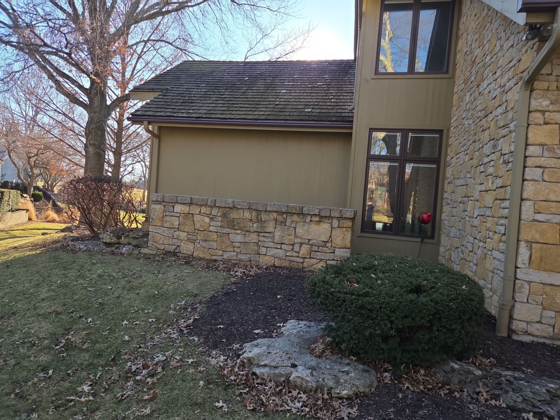 Exterior view of a two-story house with stone and tan siding, a low stone wall, and some greenery.