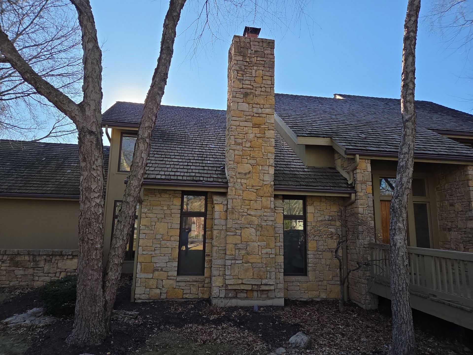 Stone chimney on a house with a gray roof, trees in the foreground, blue sky.