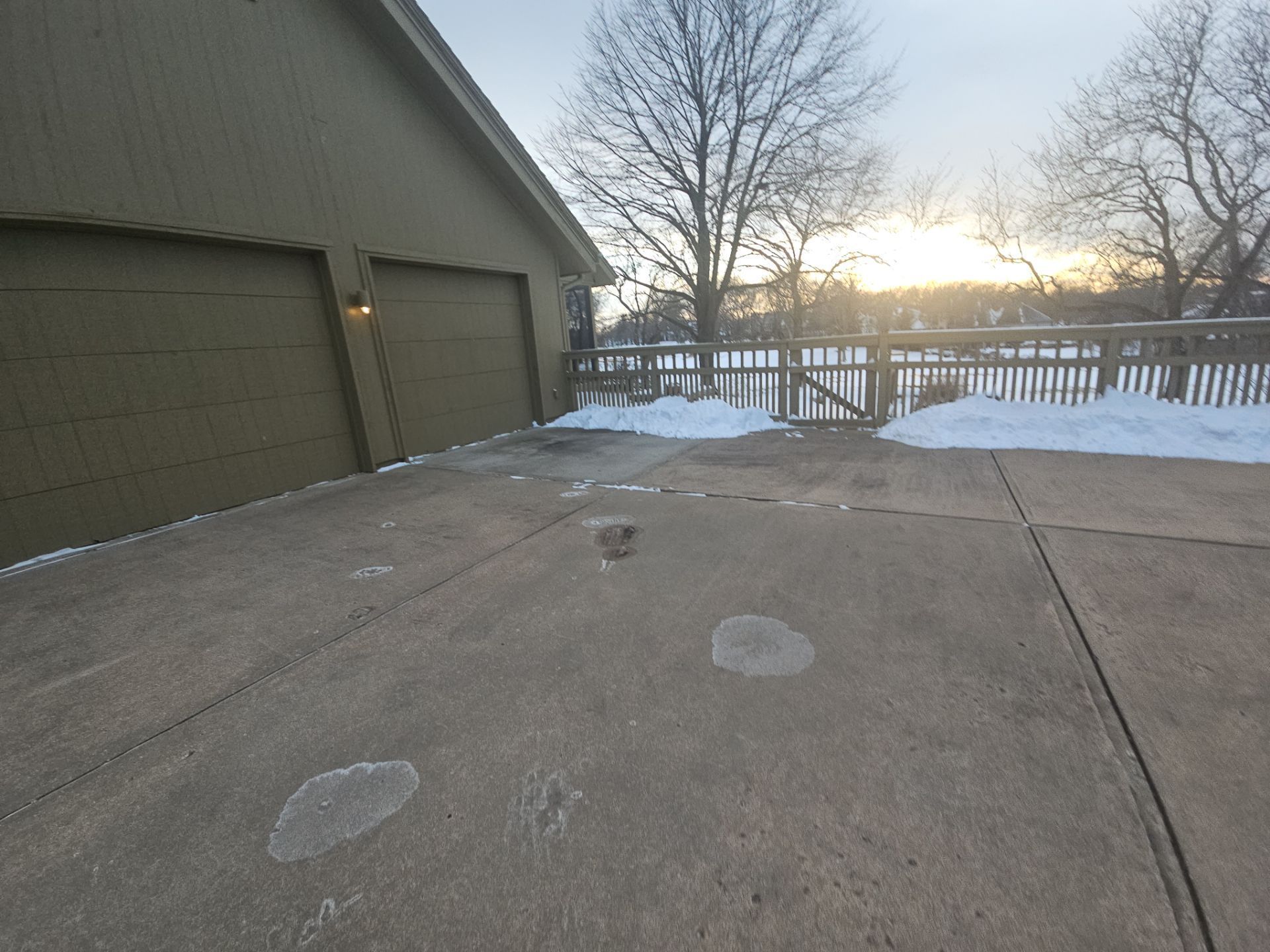 Driveway with patches of ice and snow near a garage, leading to a fenced deck overlooking a body of water at sunset.