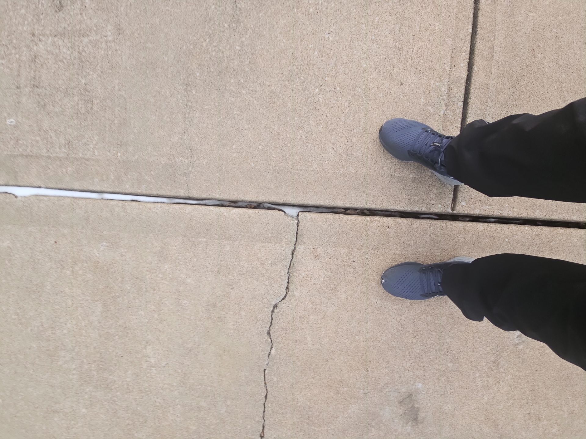 Person wearing blue boots and black pants standing on a cracked concrete sidewalk.