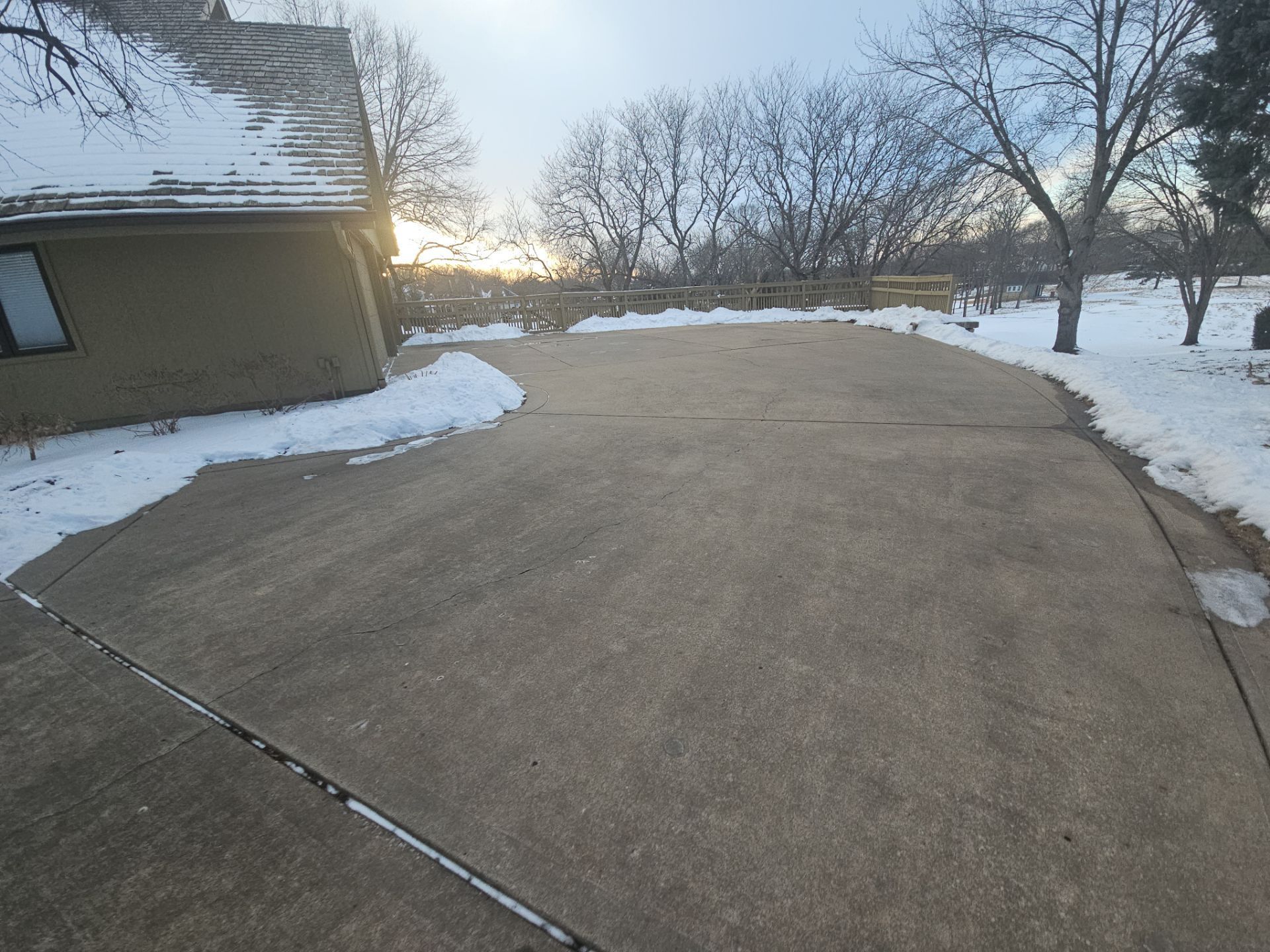 Driveway partially covered in snow, next to a house and trees on a winter day.