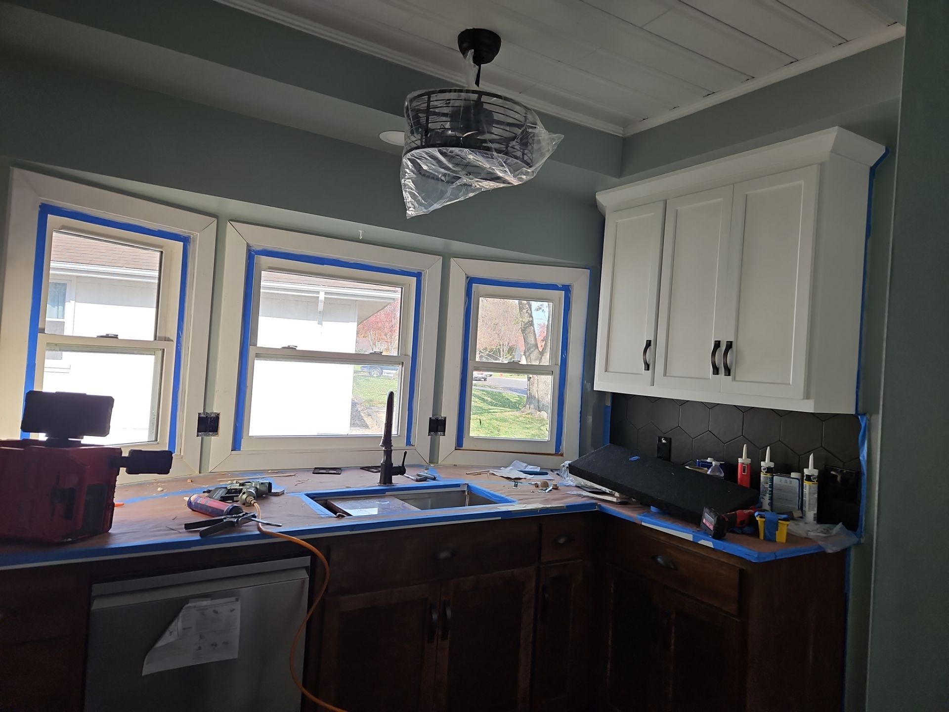Kitchen undergoing renovation: white cabinets, sink, windows taped with blue, dark wood lowers.