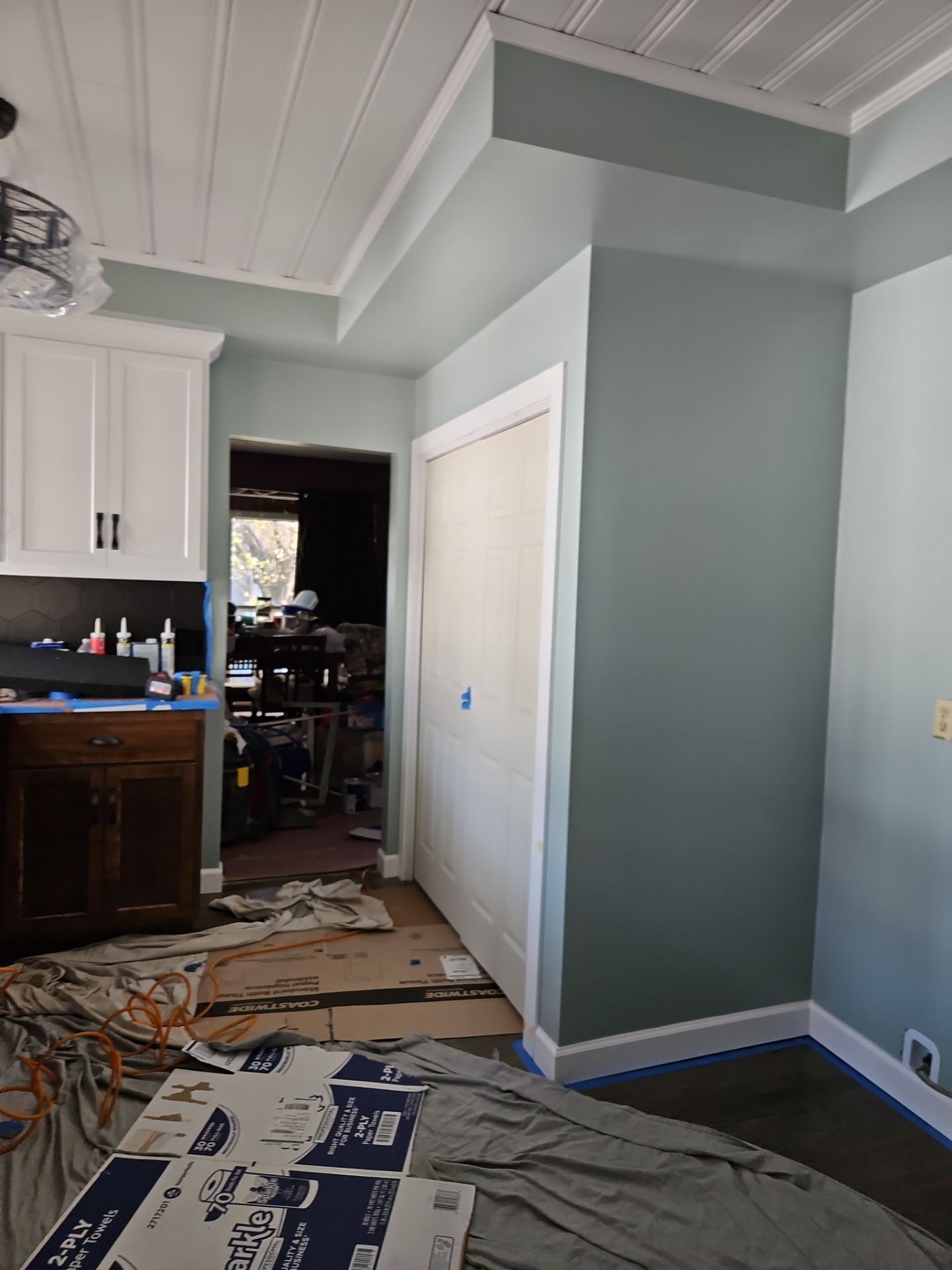 Kitchen and closet interior: white cabinets, green-gray walls, white ceiling, partially covered floor, and open doorway.