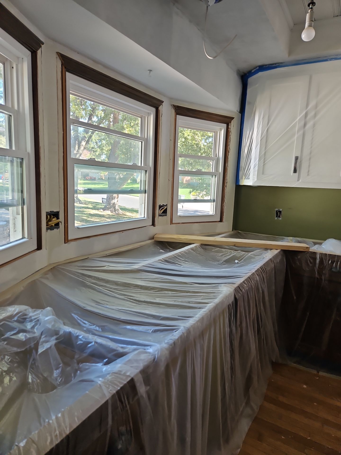 Kitchen renovation in progress; countertops and cabinets covered in plastic. White windows, brown trim, green wall.
