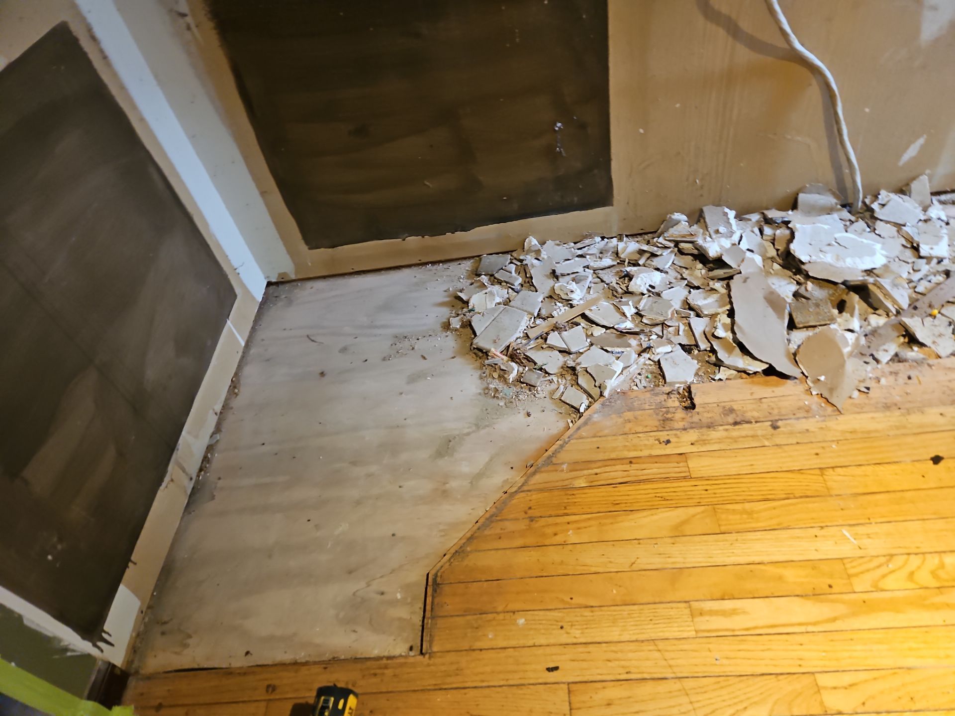Room with damaged drywall, plywood, and hardwood flooring. Debris scattered.