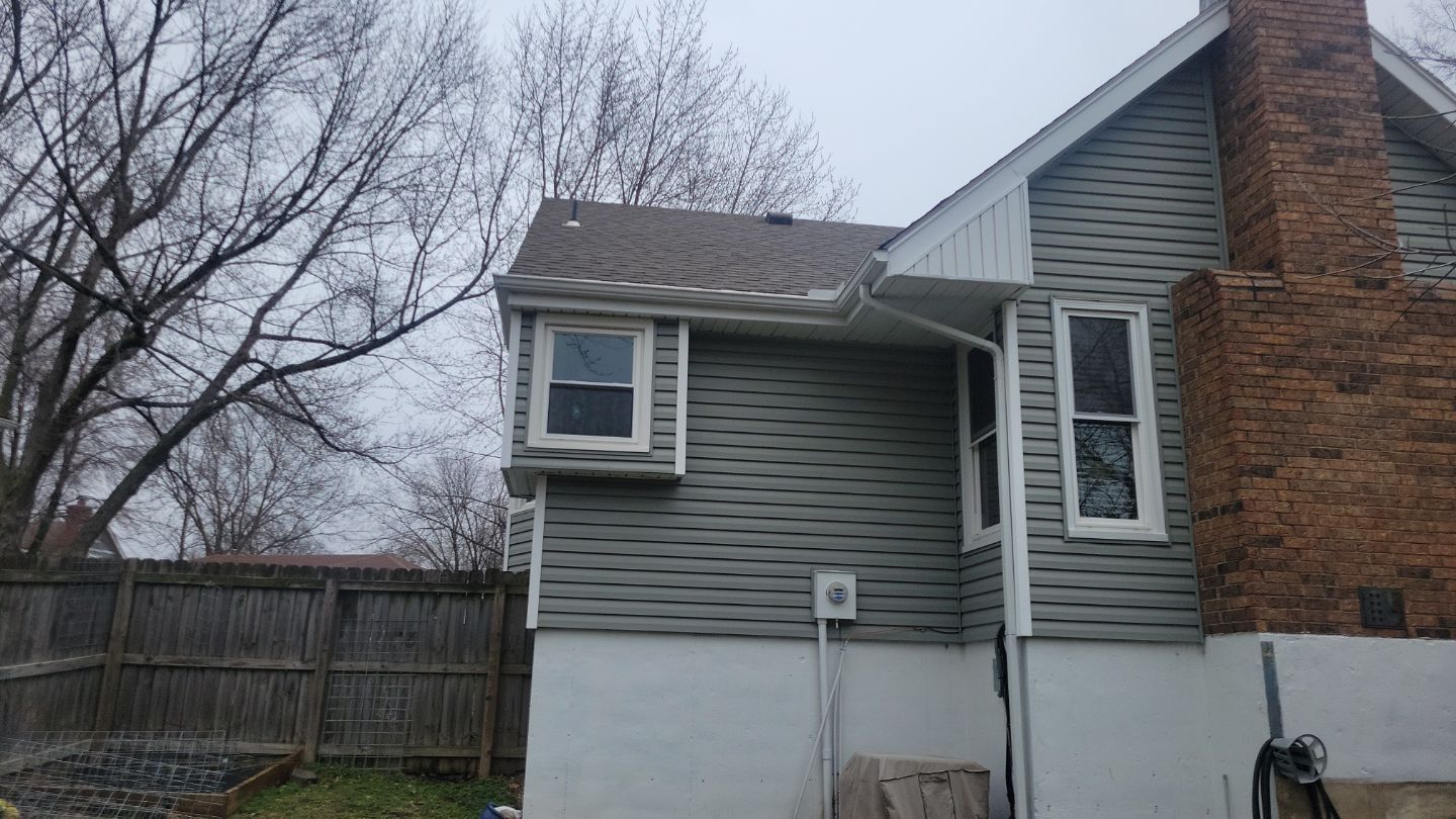 Gray house exterior with two windows, brick chimney, and a wooden fence. Overcast sky.