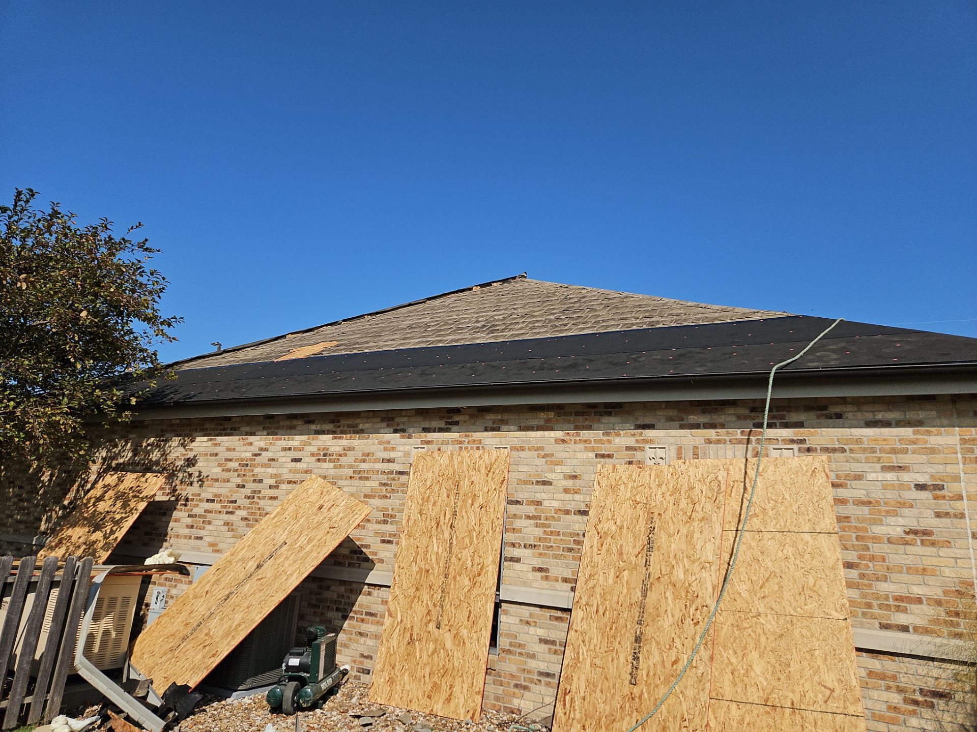 Exterior of a brick home with plywood boards leaning against the wall. The roof has shingles. Bright blue sky.