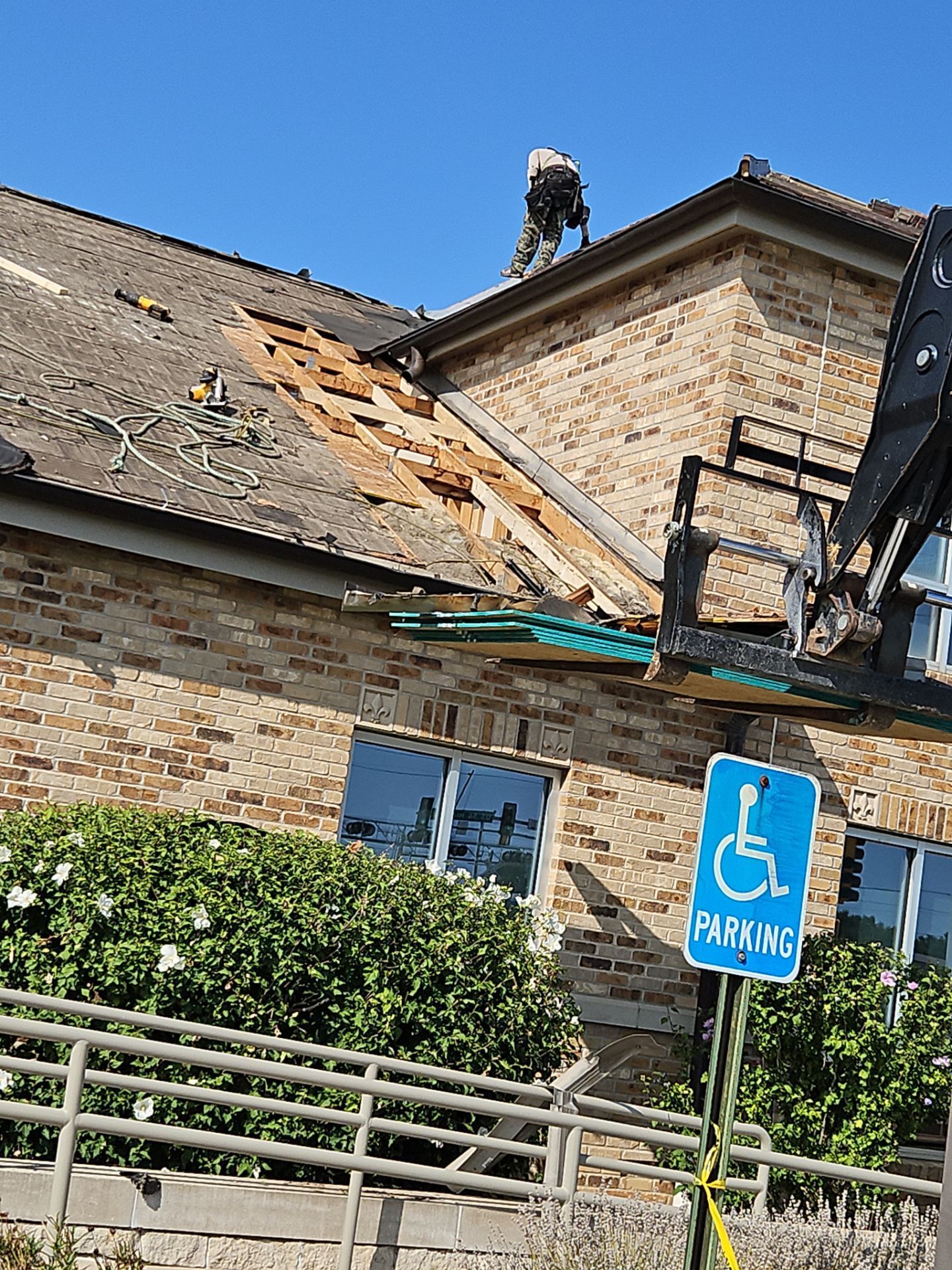 Building roof under repair; worker on roof, forklift nearby. Blue handicap parking sign in front.