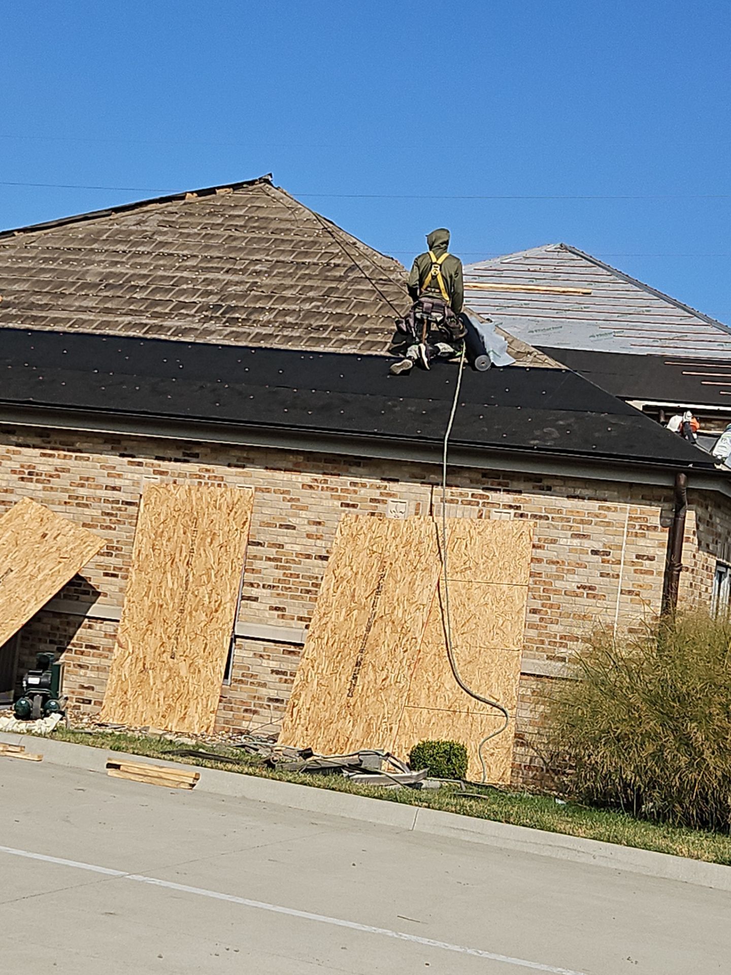 Roofer on a dark-shingled roof, near boarded-up windows and a building with a brick facade, clear sky.