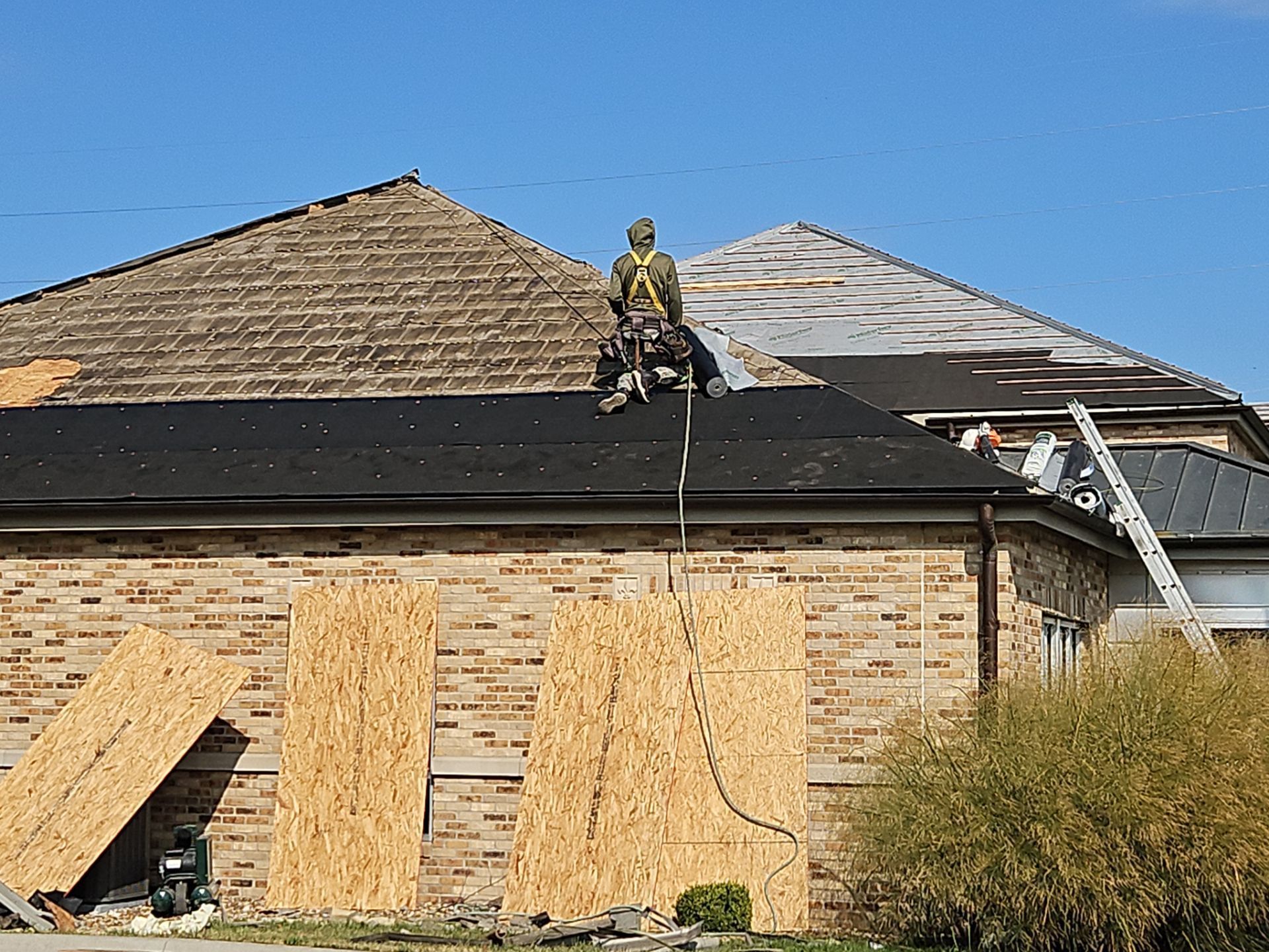 Roofer in safety harness working on a partially shingled roof; boarded up house windows.