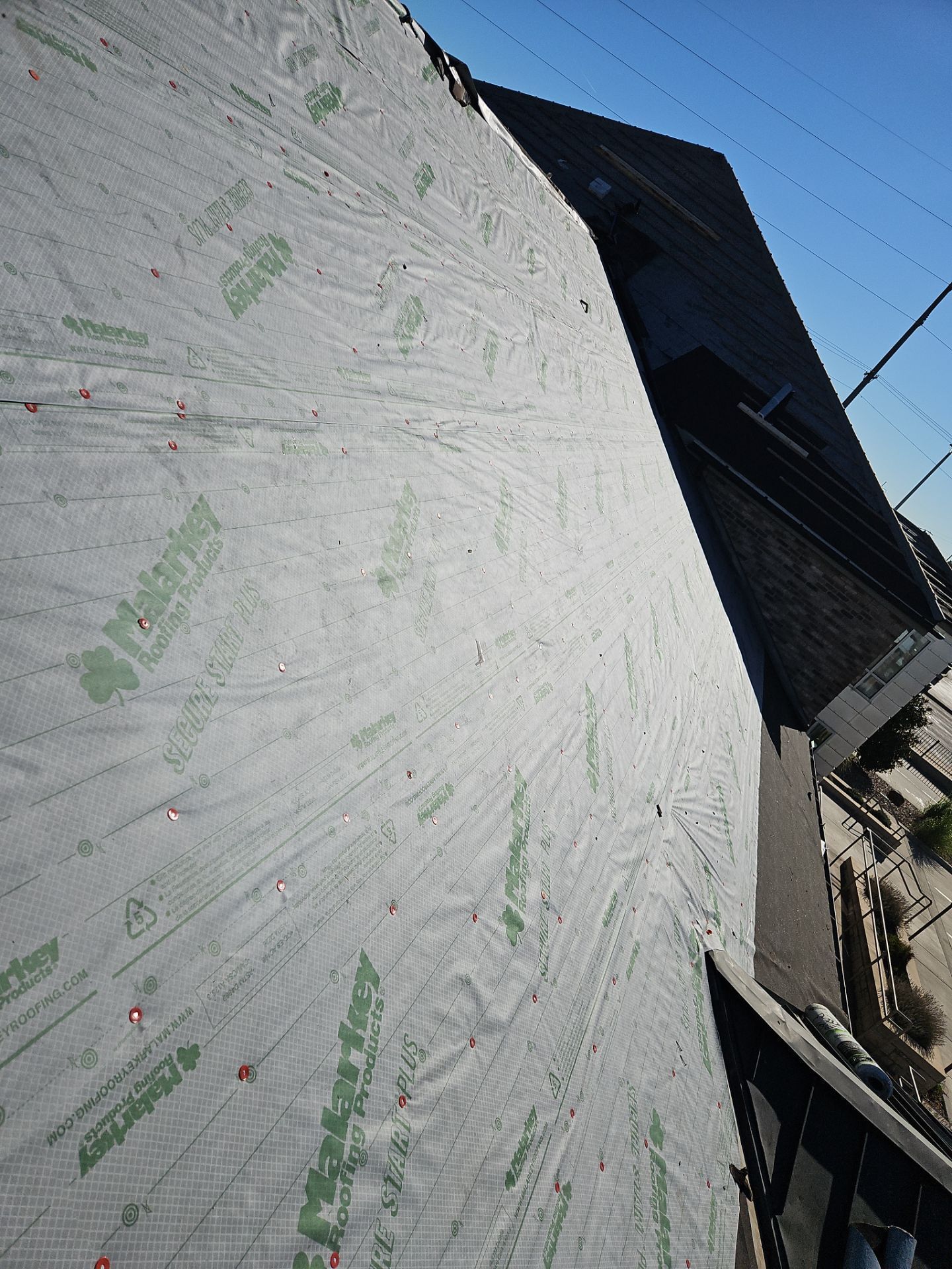 Gray roofing underlayment on a roof, with green text visible. Sky and building in background.