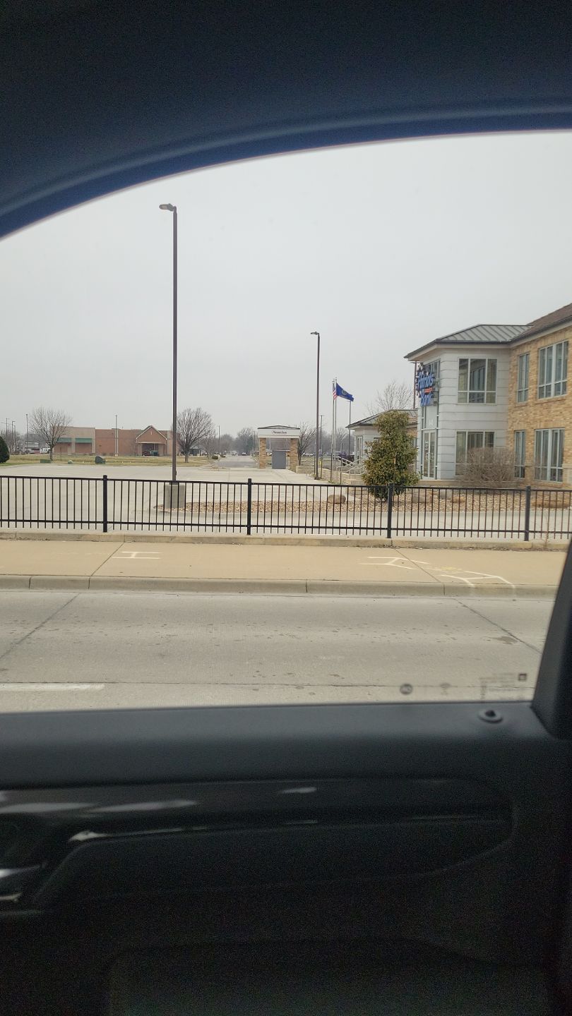 View from a car window of a street with a building, fence, and flags on a gray, overcast day.
