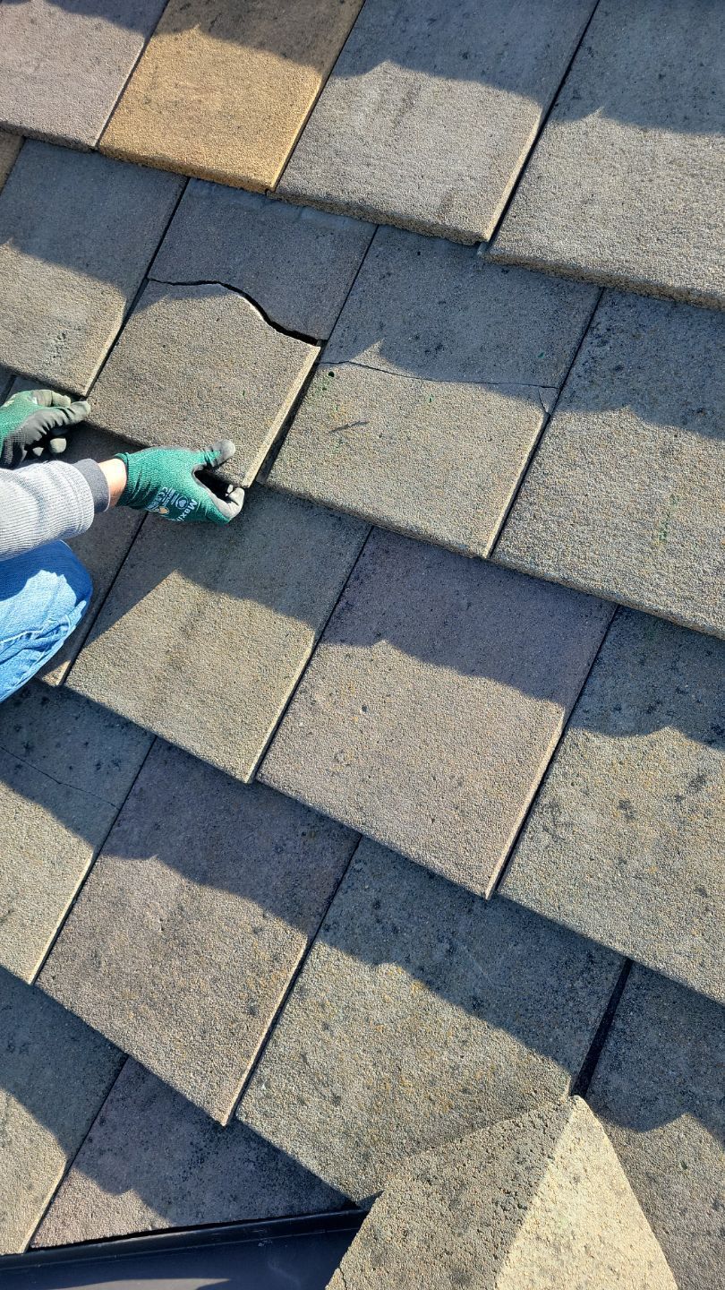 Person with green gloves examines cracked roof tile on a gray shingle roof.