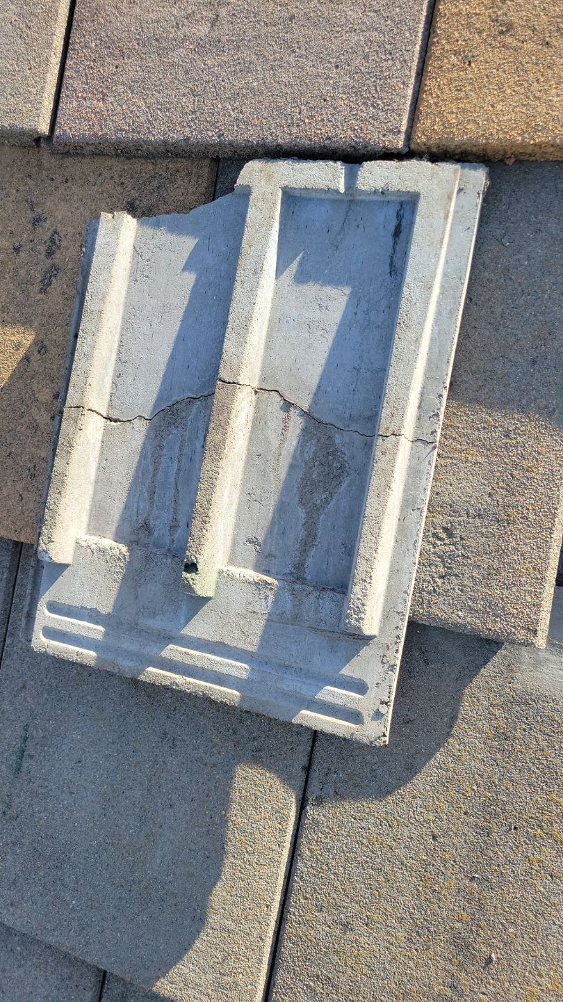 Broken concrete roof tile on an asphalt shingle roof, showing cracks and the tile's form.