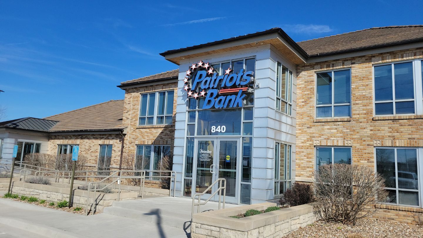 Patriots Bank building with a blue sign against a clear sky. Beige brick facade with windows.