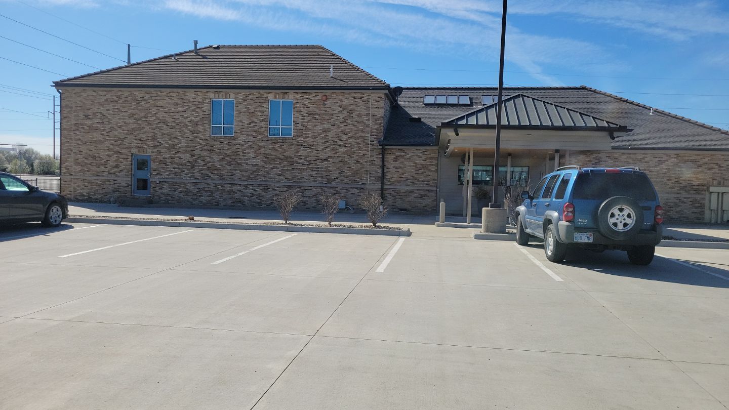 A tan brick building with blue SUV parked in front; sunny parking lot.