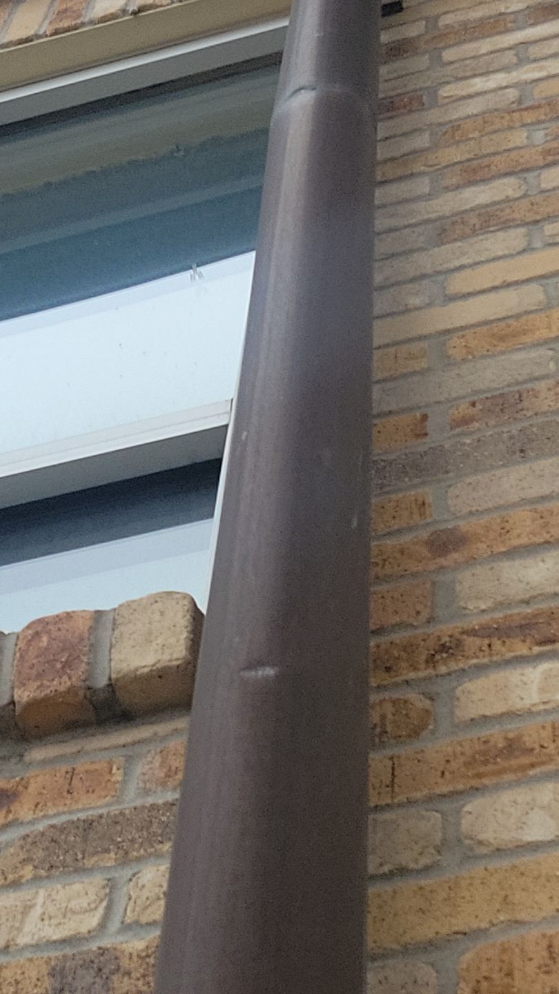 Brown downspout on a brick wall next to a white-framed window.
