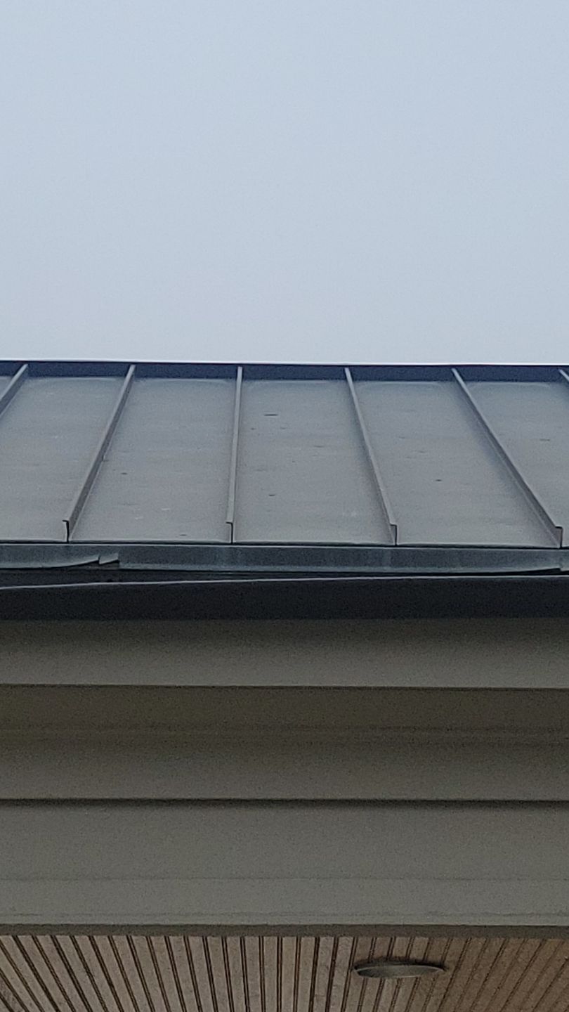 Close-up of a gray metal roof with vertical seams. A tan building's siding is below.