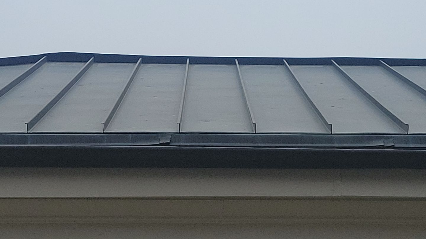Gray metal roof with raised seams over a light-colored wall.