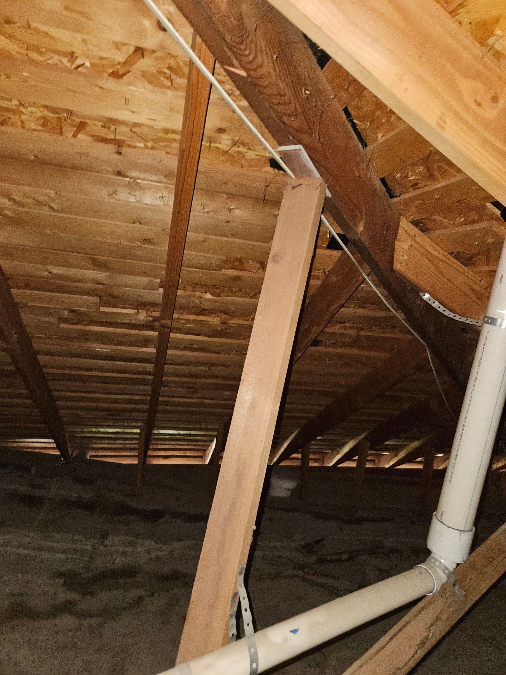 Brown attic interior with wood rafters, support beam, and plumbing pipes.