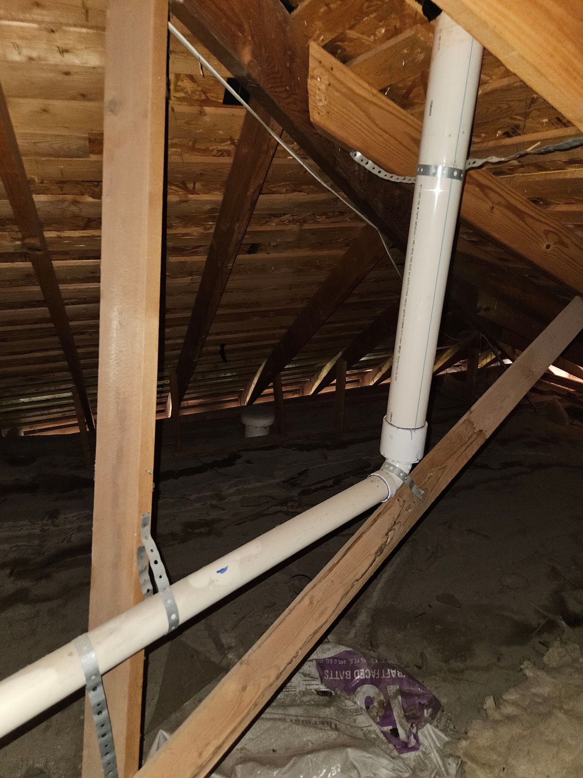 PVC plumbing pipes in an attic, connected and secured with metal straps, running along wooden beams.