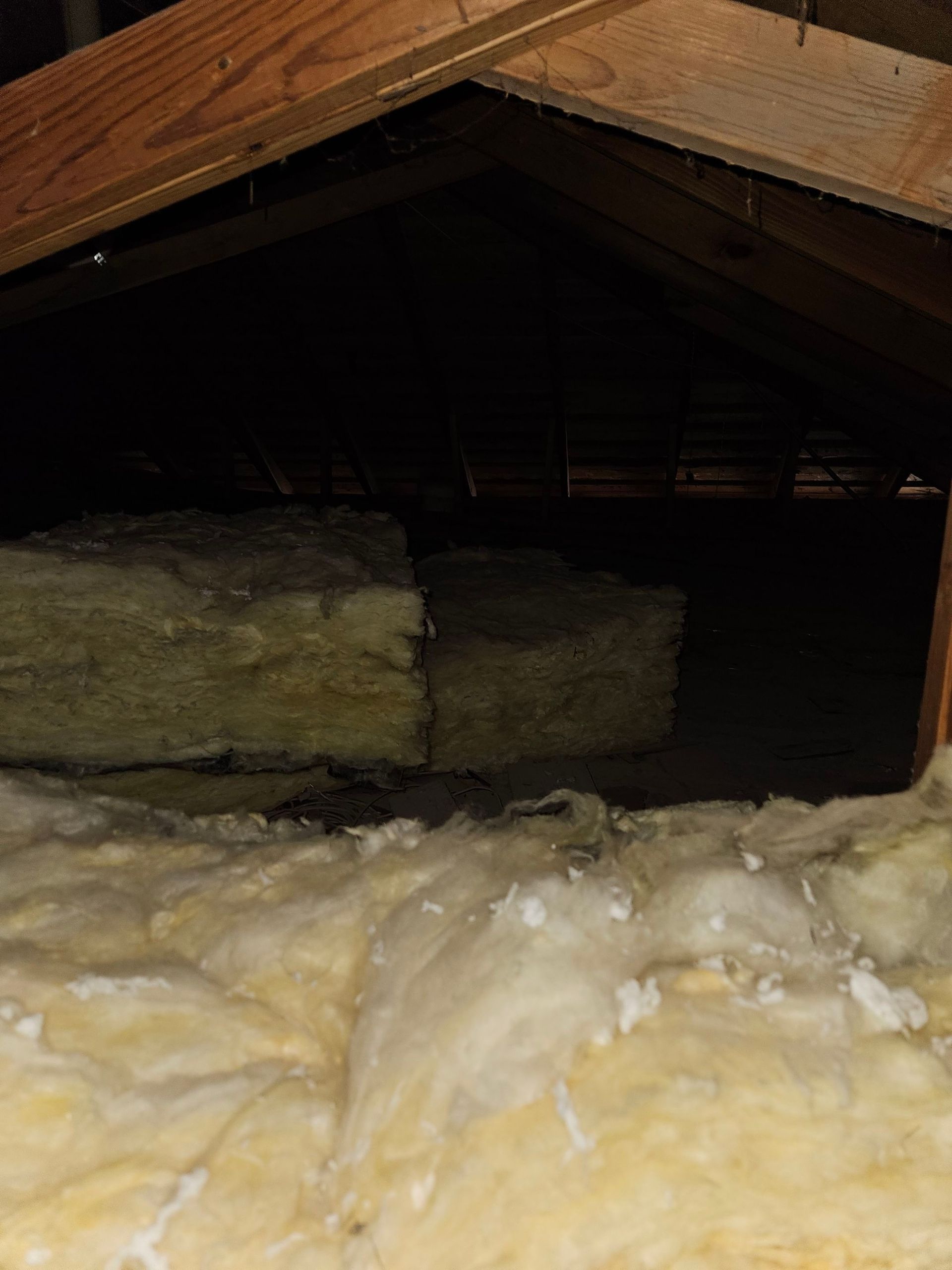 Attic interior with insulation. Beige insulation on the floor and stacked against a dark space. Wooden beams visible.